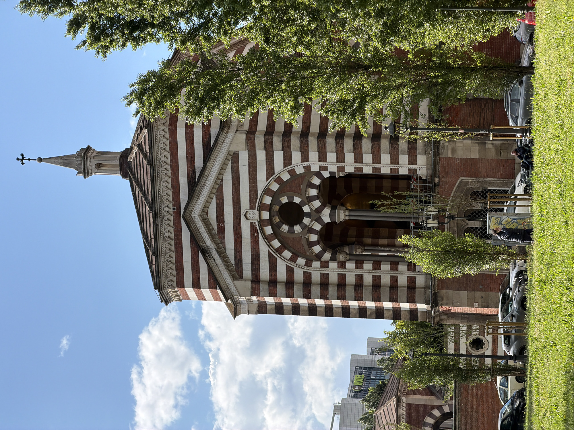 The image depicts a church with distinctive red and white striped architecture, likely a Lombard Romanesque style. The building features a central entrance with a patterned arch above, topped with a small, ornate spire. A section of the church, built with red bricks, is also visible, contrasting with the striped pattern. The building is partially obscured by trees, adding to the visual depth. In the foreground, cars are parked along the roadside, and a small grassy area is visible. A man is sitting by a street sign next to the parked cars. The sky is blue with scattered clouds, suggesting a bright, sunny day. Modern buildings appear in the background. The overall scene conveys an urban environment where historical architecture is juxtaposed with contemporary life. The location is in an unknown city.