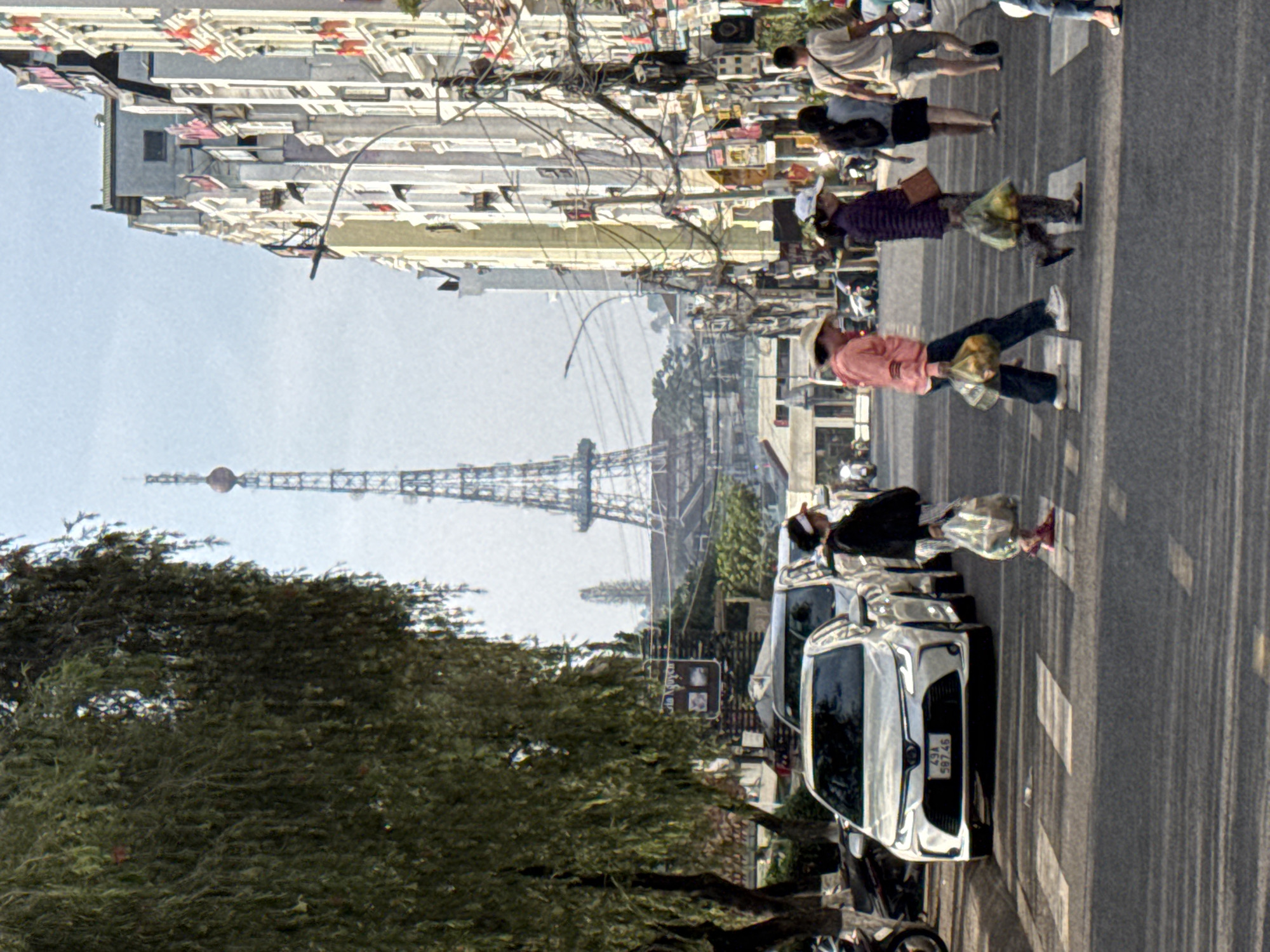 The image depicts a bustling street scene in an urban environment, likely during the daytime under clear skies. The perspective is slightly elevated, looking down a street lined with buildings and trees, with a distinctive tower in the distance.
In the foreground, an asphalt road with prominent white crosswalk markings is visible. Several pedestrians are crossing or walking alongside the street. One person, wearing a light pink shirt and a straw hat, is mid-stride on the crosswalk, carrying two plastic bags filled with items. Another individual, further back, wears a purple top and also carries bags while walking. Other people are visible further down the street and near parked vehicles.
A white car, appearing to be a Toyota sedan, is parked or slowly moving on the right side of the road, displaying a license plate with the characters "29A 597.46". Several motorbikes are also parked along the curb. Above the street, a dense network of overhead wires and cables is visible, characteristic of many urban areas in Vietnam.
To the left, a large, lush green tree with dense foliage occupies a significant part of the frame, casting shadows. In the midground and background, multi-story buildings with light-colored facades and numerous windows line the street. Dominating the background against the bright sky is a tall, slender metal-latticed tower topped with a sphere, which resembles the Da Lat TV Tower or Radio Tower.
A small street sign or advertisement is partially visible near the trees, showing the text "NGUYEN C", likely part of a Vietnamese street name. The scene conveys a sense of daily activity and movement in a lively city street.