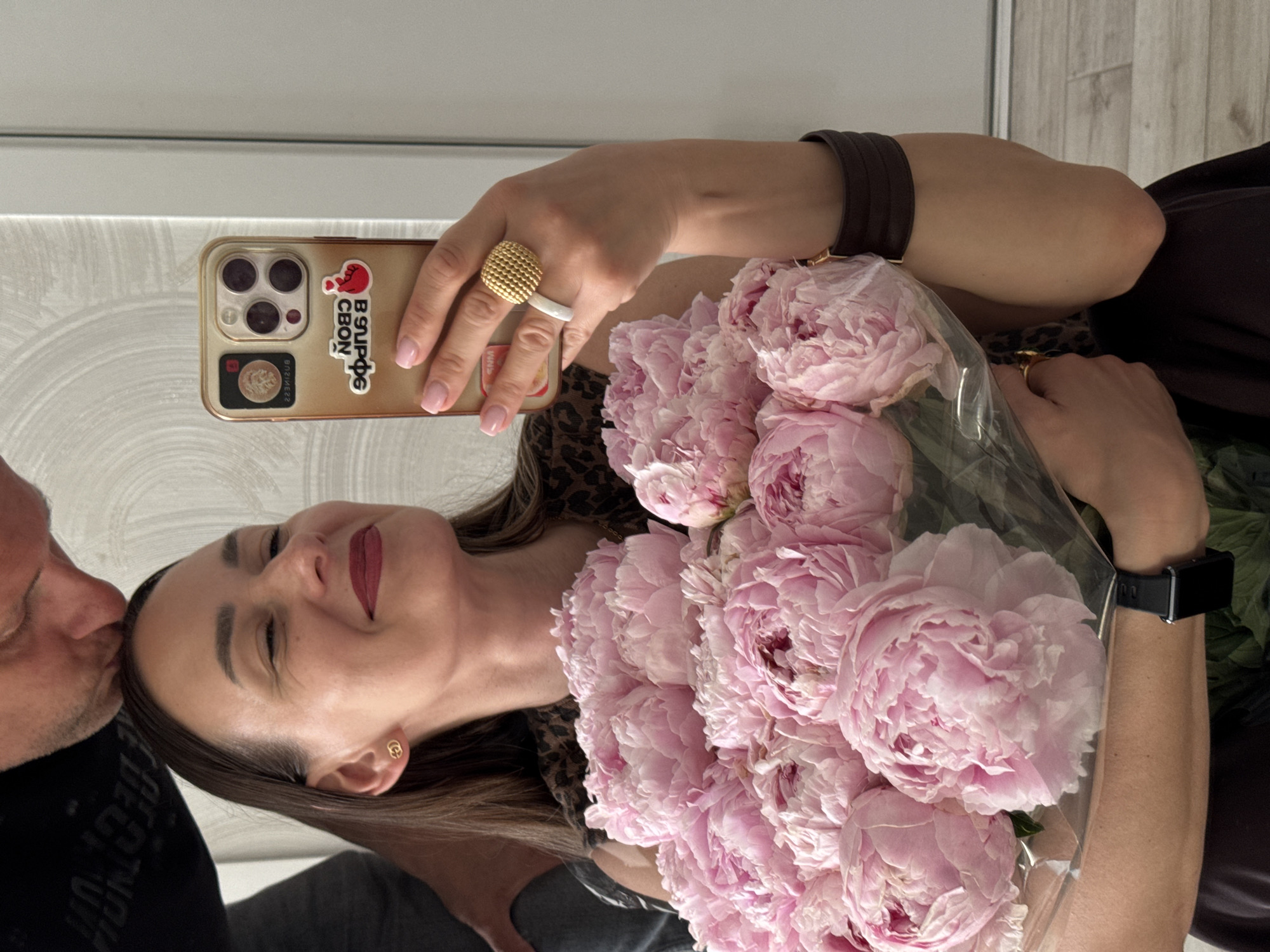 A woman is taking a mirror selfie while a man kisses her forehead. She is holding a large bouquet of pink peonies and is wearing a leopard print top, a wide brown cuff bracelet, and a gold ring. The man is wearing a black t-shirt with white lettering. The background appears to be an indoor setting with light-colored walls and a wooden floor. The image does not provide any specific details that would indicate the location is in Sochi, Russia.