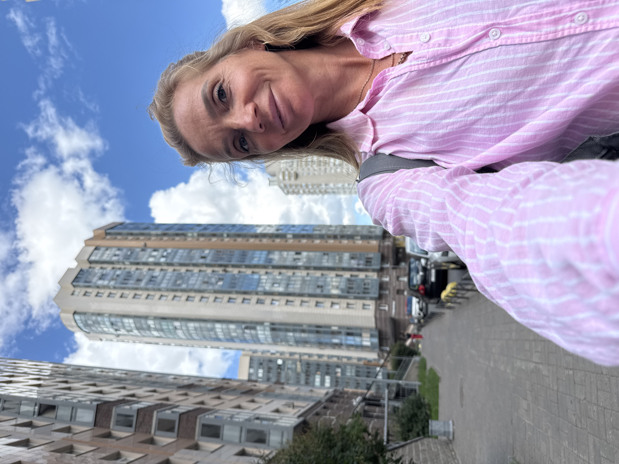 A woman with blonde hair, wearing a pink and white striped shirt and a backpack, takes a selfie on a sidewalk in front of tall buildings. The sidewalk is paved with gray bricks and there are parked cars on the street to the right. The sky is blue with white clouds. The buildings appear to be apartment complexes. This scene is likely located in Vsevolozhsk, Russia.