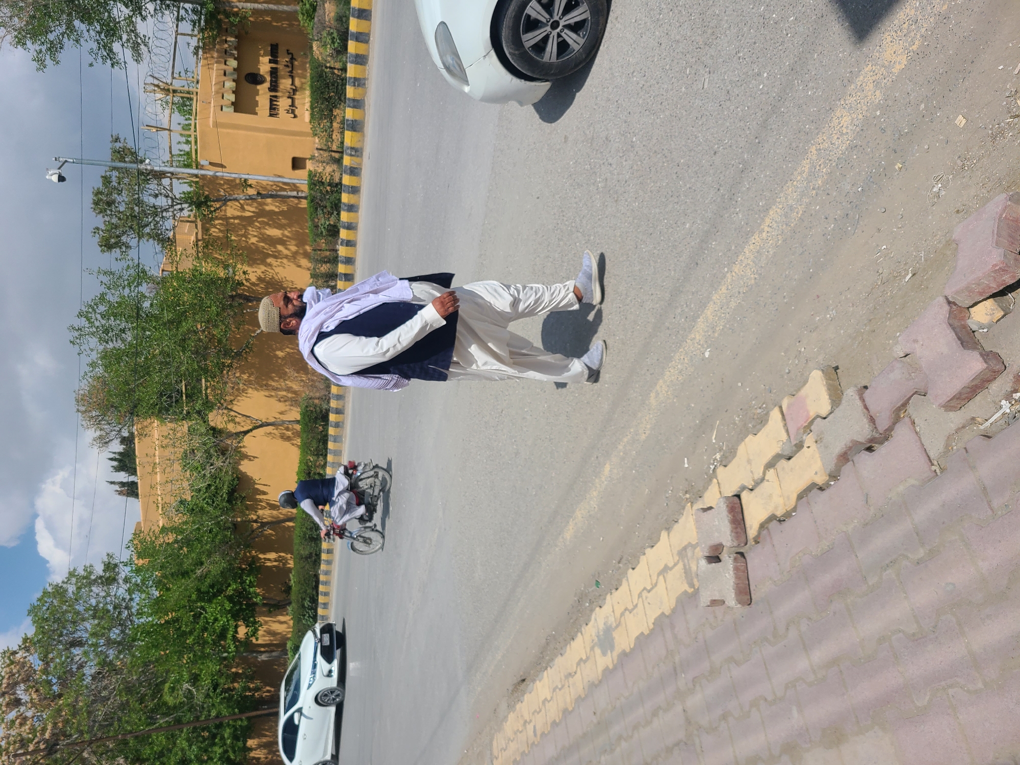 A man in traditional Pakistani clothing, including a white shalwar kameez, a dark waistcoat, and a white turban-like head covering, is walking along a street in Quetta City Tehsil, Pakistan. He is walking towards the right side of the image. A white car is parked on the street near him, and another white car is further down the road. In the background, there is a building with yellow and black striped markings along the road in front of it, suggesting a curb or barrier. A man is riding a bicycle on the road in the background. The road is asphalt. The sidewalk consists of red and yellow bricks. The sky is visible with some clouds. Trees are alongside the road.