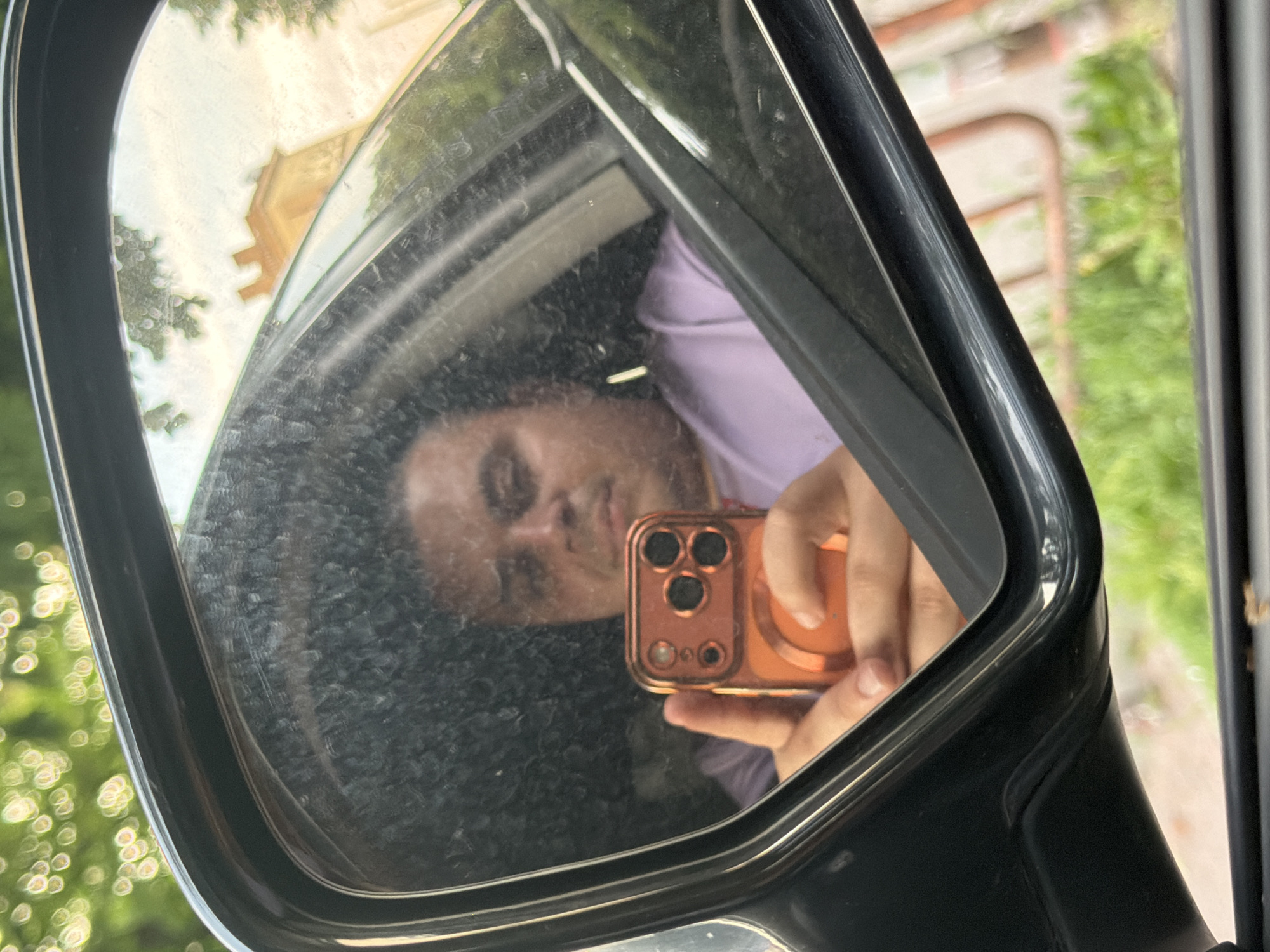 The image captures a selfie taken through a car's dusty side mirror. The reflection shows a person, likely the photographer, looking directly towards the smartphone they are holding. The person has dark hair and is wearing a light purple or lavender top.
The smartphone, held in the person's hand, is orange or copper-colored and features a distinctive three-lens camera module. The mirror's surface is noticeably smudged and dirty, especially around the reflection of the person's face and shirt.
The background reflected in the mirror reveals an outdoor daytime setting. Out-of-focus green foliage and trees are visible in the upper left, while a light-colored building or structure with a geometric roofline can be seen faintly in the upper central portion of the reflection. The dark frame of the car's side mirror borders the reflective surface. There is no visible text in the image. The general setting suggests an outdoor location, with the given context of Shumen, Bulgaria.