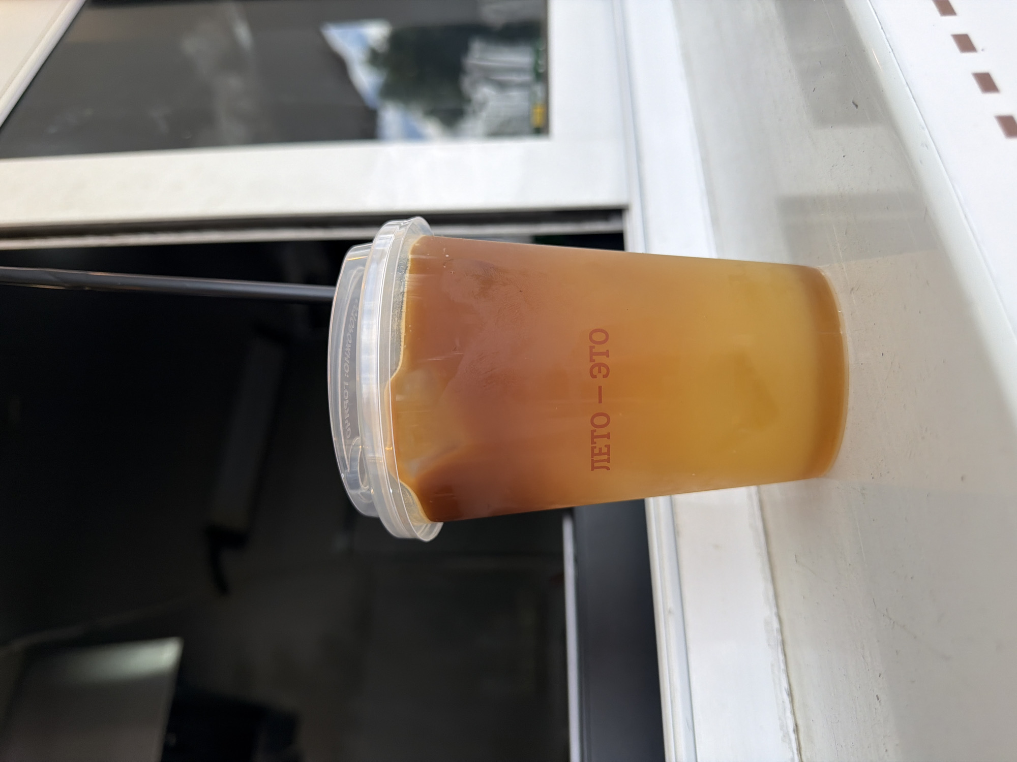 A close-up, eye-level, medium shot captures a clear plastic cup with a translucent lid filled with a layered beverage. The drink displays a gradient of color, with a darker amber liquid settling on top of a lighter yellow liquid. A subtle red Cyrillic inscription, "ЛЕТО – ЭТО", is visible on the lower portion of the cup. The cup is placed on a white, slightly textured surface, possibly a windowsill or counter, with a soft shadow cast to its right. In the background, out of focus, is a dark environment with vertical elements, suggesting an interior space or a shaded area, and a bright reflection in a window or glass surface, showing a blurred image of trees and a sky, hinting at an outdoor scene beyond the immediate location. The overall impression is a casual, potentially takeaway beverage. The inscription "ЛЕТО – ЭТО" translates to "Summer is this" in Russian, a phrase commonly associated with seasonal offerings, particularly ice cream or cold drinks, and is reminiscent of branding found in cafe or kiosk establishments in Saint Petersburg, Russia.