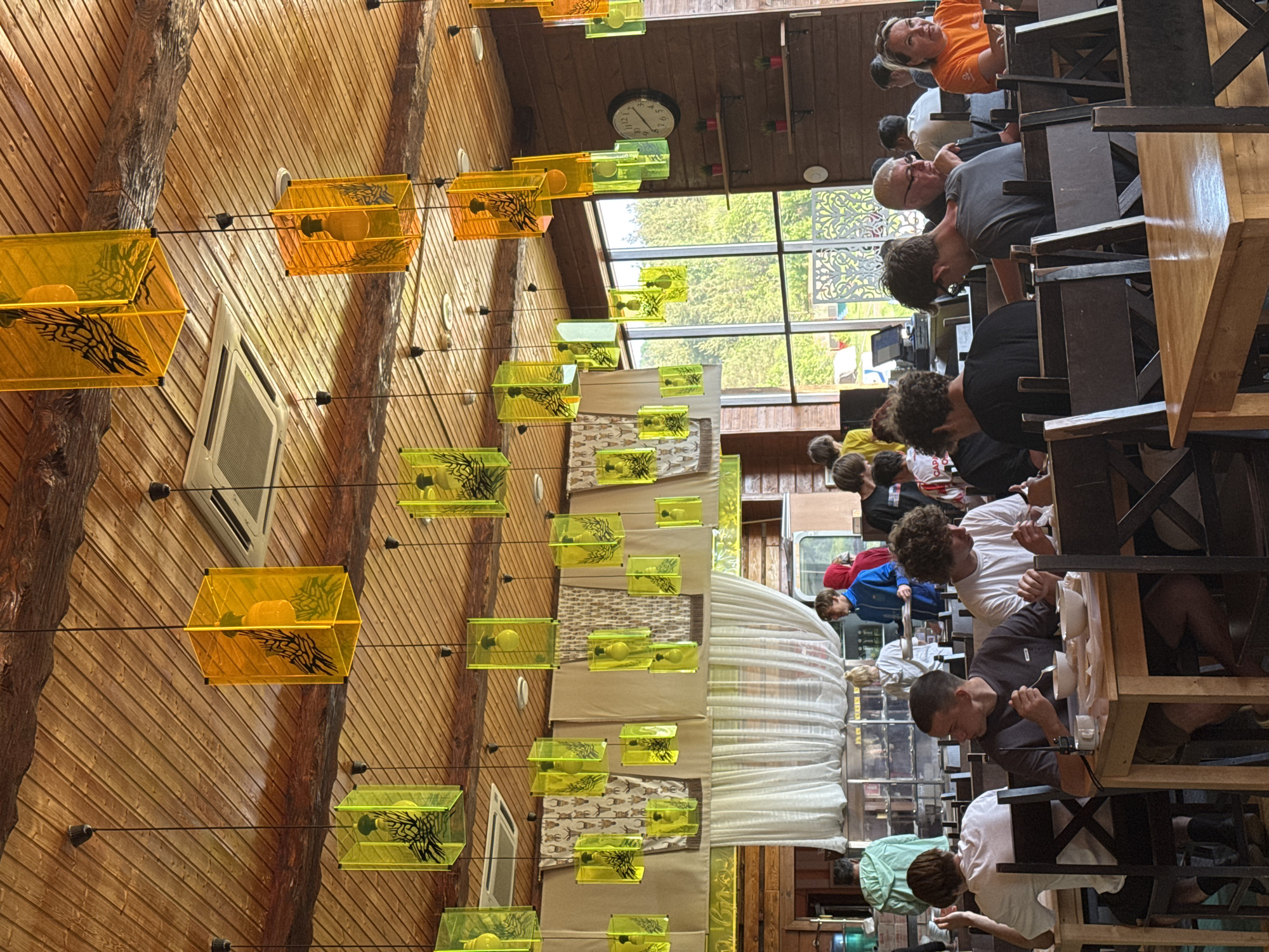 The image shows the interior of a restaurant in Khvalynsk, Russia. The restaurant features unique decor, including rows of square-shaped pendant lights in yellow and green hues, suspended from the ceiling. The walls appear to be paneled with wood, interspersed with what seems to be tree trunks or large branches as design elements. There is a clock visible on the wall towards the back of the room, and a window providing natural light. Tables are arranged in rows, and many people are seated, eating and socializing.