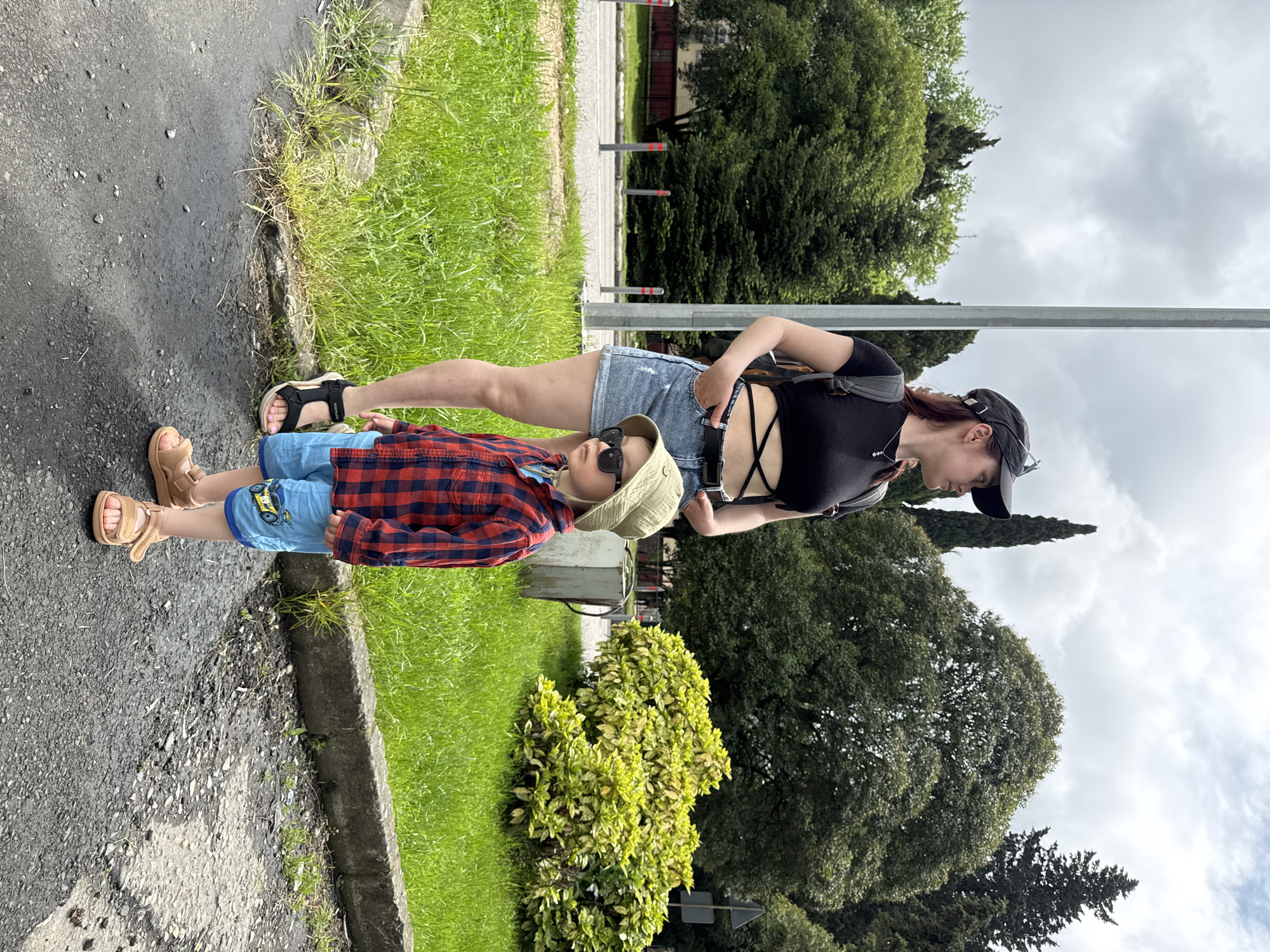 A woman and a young child stand next to a road. The woman wears a black top, denim shorts, a backpack, and a black baseball cap, and sandals. The child wears a red and blue plaid shirt, denim shorts with a monster truck graphic, a hat, sunglasses, and brown sandals. They stand next to a road with a curb, a grassy area, a shrub, and trees in the background. The location is in Sochi, Russia, judging by the roadside posts and foliage in the background. The sky is overcast.
