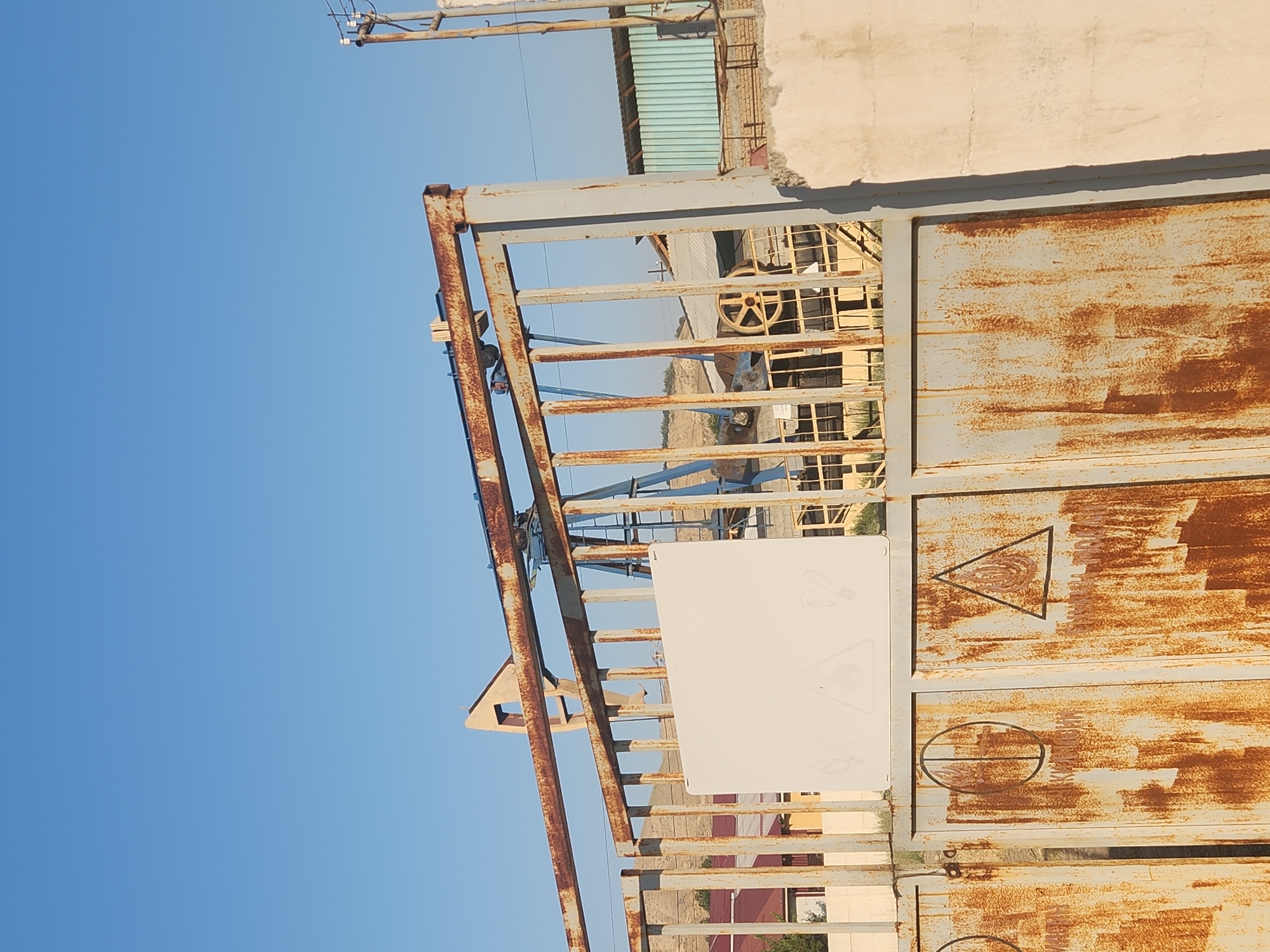 The image captures an outdoor scene dominated by a tall, rusted metal gate. Behind the gate, a portion of an oil pump jack is visible against a clear blue sky. The gate itself features two large, weathered panels with visible rust and some markings. One panel on the left shows a circular symbol with vertical lines, and the panel on the right displays a triangular symbol with an internal flame-like graphic. A blank white sign is affixed to the upper section of the gate. In the background, there are several industrial-looking structures with corrugated metal roofs and brickwork, as well as some distant hills. The overall impression is one of an industrial or operational site in a dry, possibly arid environment. The location is identified as Xidirsho, Uzbekistan.
