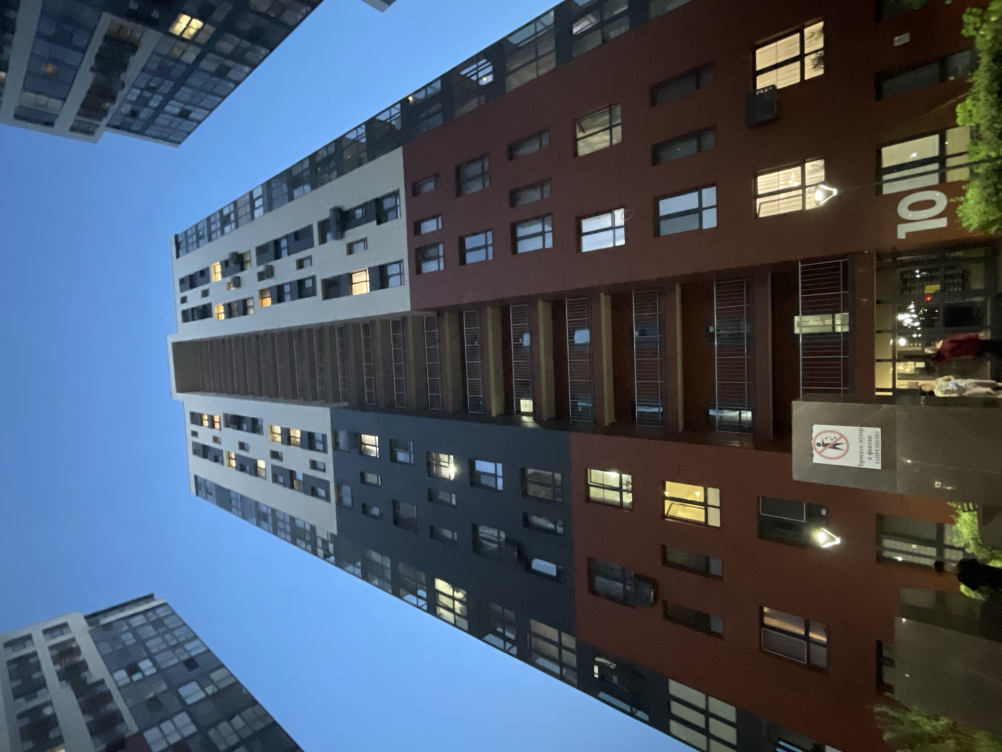 The image captures a low-angle view of a modern, multi-story residential building at dusk in Almaty, Kazakhstan. The building features a distinctive color-blocked facade with sections of dark gray, white, and maroon. Numerous windows, some illuminated from within, dot the structure. Balconies with metal railings are visible on several floors. In the foreground, a street lamp stands illuminated, and a sign with a prohibition symbol and Russian text is mounted on a dark stone structure. Two individuals are partially visible near the entrance of the building. To the left and right of the central building, other tall residential buildings are partially visible against the twilight sky. The overall environment appears to be a contemporary urban residential complex.