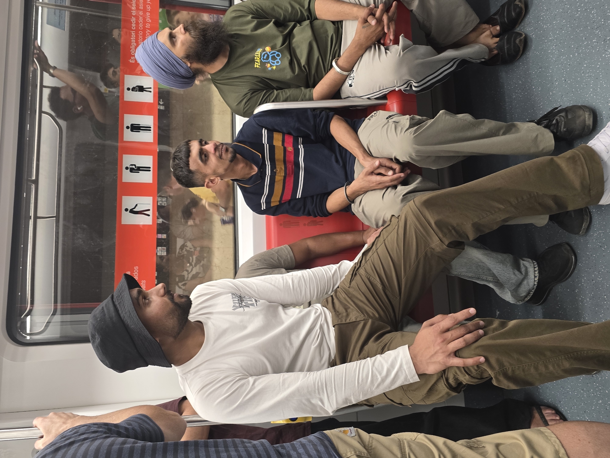 The image shows the interior of a public transport vehicle, possibly a subway or train, with several individuals seated. The setting appears to be in Cornellà De Llobregat, Spain, as indicated by signage in Spanish visible through the window.
In the foreground, three men are prominently featured. The man on the left is wearing a white long-sleeved shirt and brown cargo pants, with a black bucket hat. He is looking to his right. To his right, a man in a dark blue polo shirt with horizontal stripes of red, yellow, and white is seated, looking to his left. He has short, dark hair and is wearing light brown pants. On the far right, a man wearing a green t-shirt and a blue turban is seated, looking towards the right. His t-shirt has a graphic of a teddy bear and the word "FEARLESS" on it. He has a long, dark beard.
The seating appears to be red upholstered benches. Behind the seated individuals, there is a window revealing the outside, or possibly another part of the vehicle, where other passengers are visible. A red band with pictograms of people, including one with a cane and a pregnant woman, runs horizontally across the interior of the vehicle. This band is likely indicating priority seating or rules for passengers. The floor is a speckled gray material. The overall environment suggests a functional, urban public transportation setting.