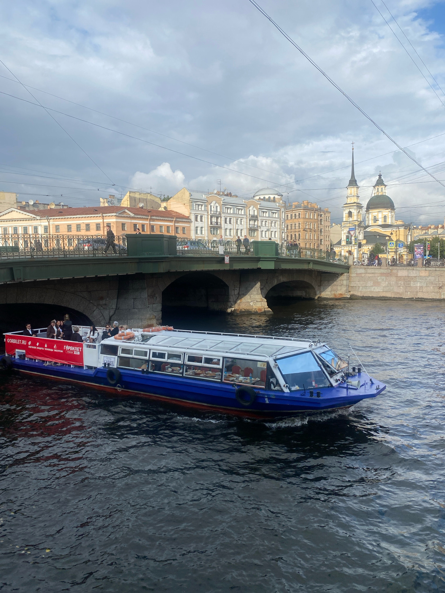 A blue and white tour boat with a red banner runs along a dark canal in Saint Petersburg, Russia. The boat is filled with passengers and has an enclosed cabin with large windows. In the background, a stone bridge spans the canal, with buildings and a yellow domed church visible beyond it. The sky is overcast with clouds.