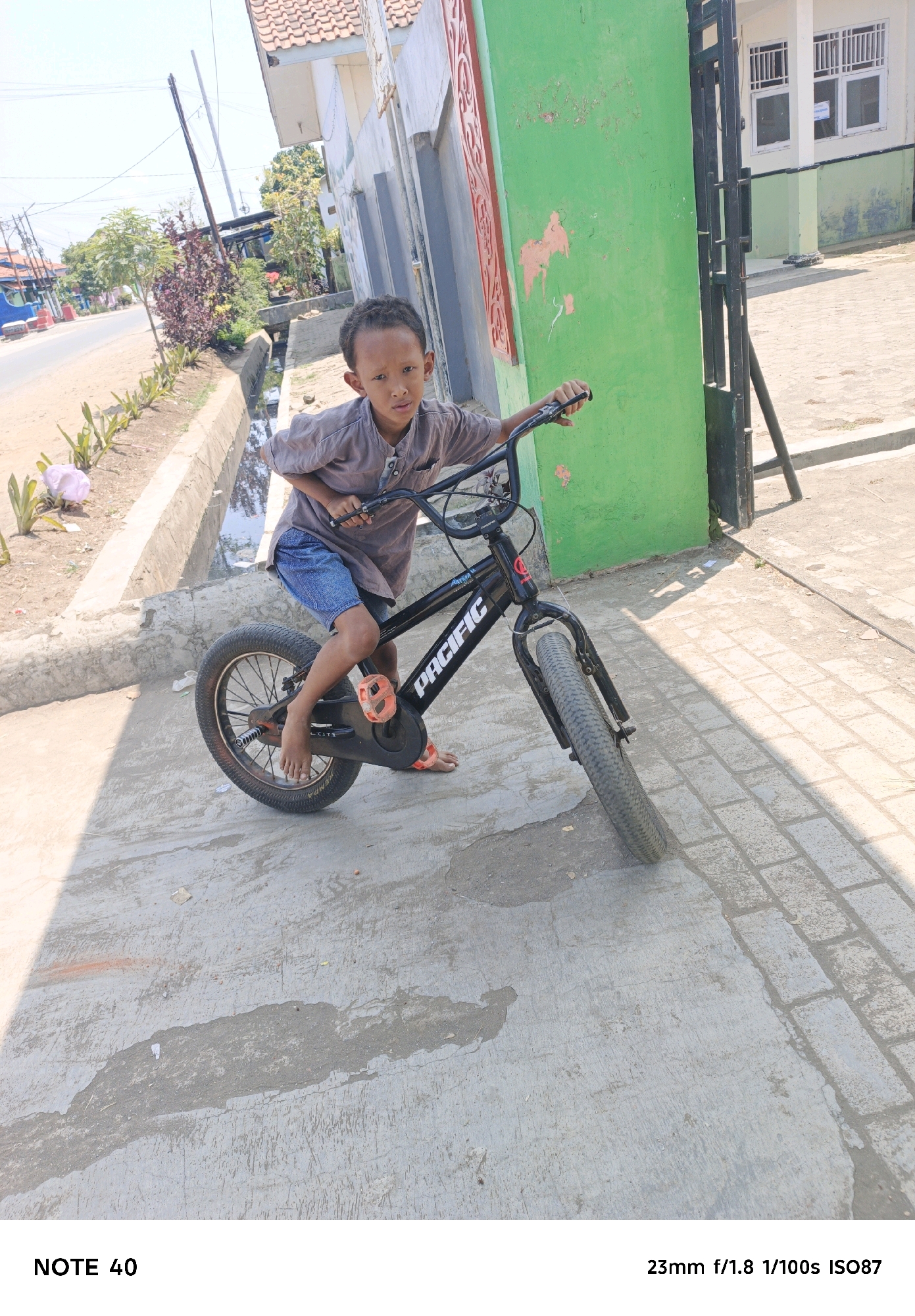 A young boy is sitting on a black bicycle with wide tires and orange pedals on a concrete sidewalk. He is wearing a gray shirt and blue shorts. The bicycle has "PACIFIC" written on the frame. The boy is looking at the camera. In the background, there is a building with green and white walls, a red decorative element, and a black metal gate. To the left of the building, there is a street with a ditch running alongside it, and some vegetation. The scene appears to be in an outdoor urban or semi-urban environment in Kelurahan Cilacap, Kecamatan Cilacap, Indonesia. The lighting suggests it is daytime with sunlight casting shadows.