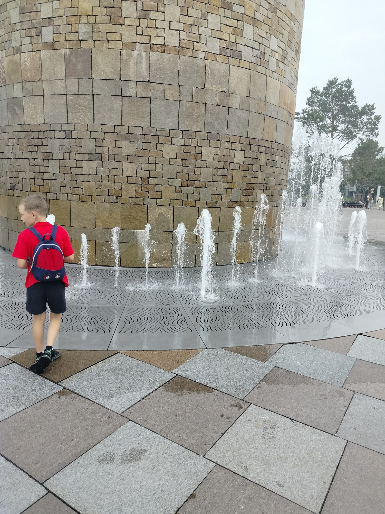 A young boy, viewed from the rear, stands on a patterned tiled surface in front of a circular stone structure and a series of water fountains. The boy is wearing a red t-shirt, black shorts, a red and blue backpack, and dark sneakers. The stone structure, made of rough-hewn rectangular blocks, forms a curved wall. Several water jets shoot upwards from the tiled ground in front of the structure. The tiles are a mix of gray and tan squares, with a patterned section near the fountains featuring swirling lines. In the background, trees and a slightly hazy sky are visible. The boy appears to be observing the fountains. The scene is likely an outdoor public space. This location is identified as being in Kemerovo, Russia.