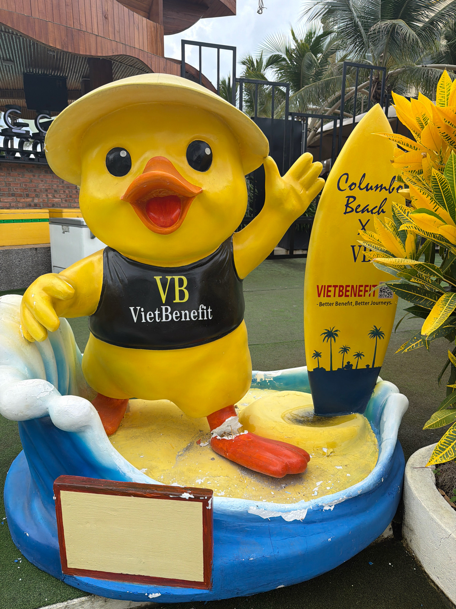 The image shows a brightly colored statue of a yellow duck wearing a hat and a black vest with "VB VietBenefit" printed on it. The duck is standing on a blue and yellow wave-like platform, with a yellow surfboard behind it that reads "Columbus Beach" and "VIETBENEFIT - Better Benefit, Better Journeys." The statue is located outdoors, with palm trees and foliage visible in the background. The overall scene suggests a beach resort or a tourist attraction in Đà Nẵng, Vietnam. The statue is detailed and eye-catching, likely designed to attract visitors.