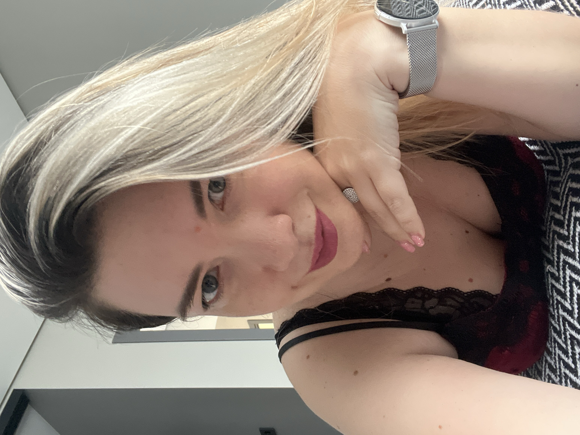 A woman with blonde hair and light skin is posing for a selfie. She is wearing a black lace top and a watch on her left wrist. Her right hand is resting on her chin, and she is wearing a ring on her right index finger. The background is blurred and appears to be indoors, with a wall and a bed visible. The image is well-lit, and the woman's face is in focus. There is no indication of the location being Sochi, Russia.