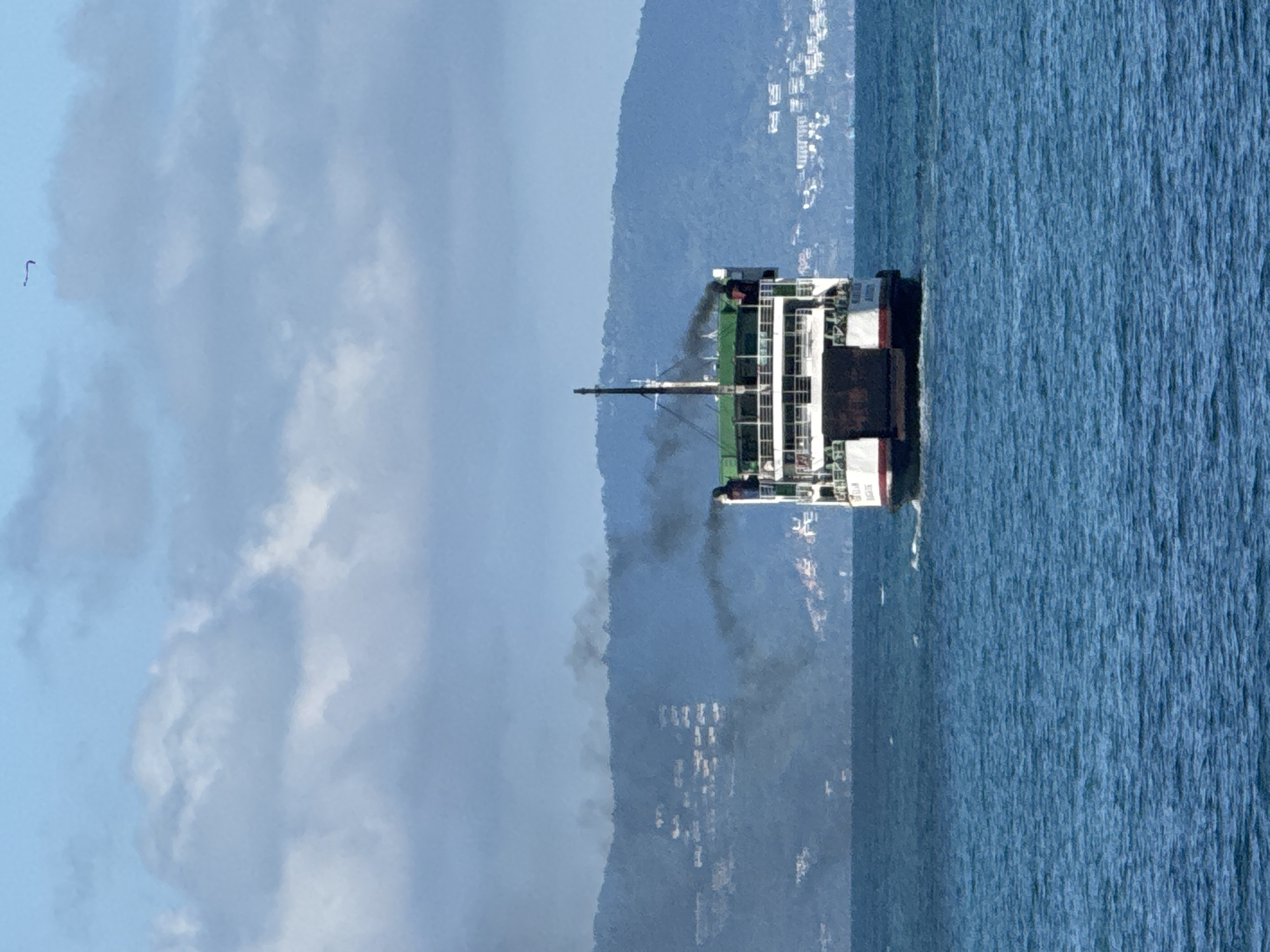 A stern view of a ferry boat is captured at sea, with dark smoke billowing from its smokestack. The ferry, painted white with green and red accents, is moving away from the camera, leaving a wake on the choppy blue water. In the background, a hazy, mountainous coastline is visible, dotted with buildings that suggest a populated area. The sky is a light blue with scattered, soft gray clouds. The location is identified as Ko Phangan, Thailand.