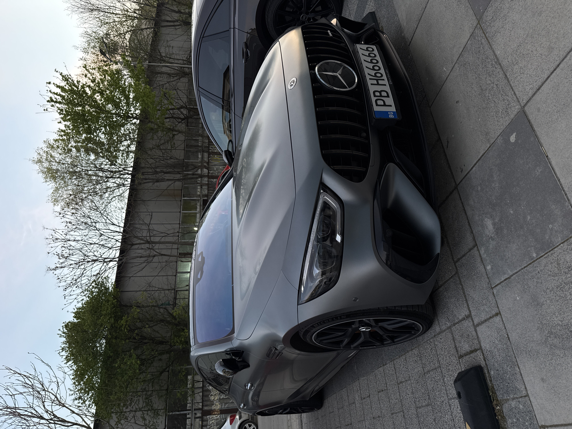 A gray Mercedes-Benz car is parked on a paved surface, likely a sidewalk or street, in an urban environment. The car's front is visible, including the grille, headlights, and license plate, which reads "PB H66666" with the "BG" country code, indicating it is registered in Bulgaria. Another dark-colored vehicle is partially visible behind it. The background features a building with windows and trees, suggesting a street scene. The location is Plovdiv, Bulgaria, based on the license plate information. There are no visible people in the scene.