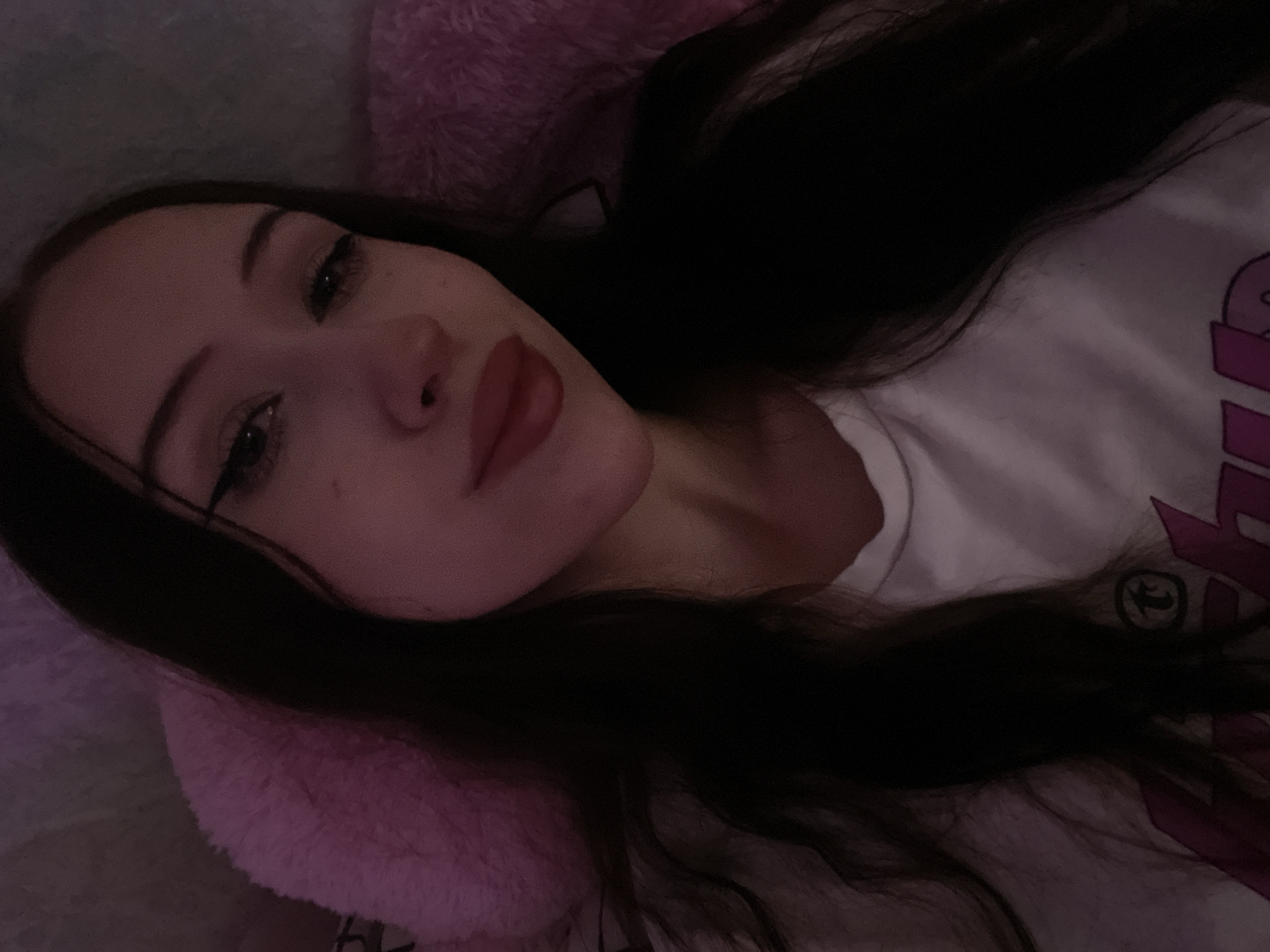 A young woman with dark hair and dark eyes, wearing a white t-shirt, poses for a selfie. She has dark eyeliner and pink lipstick. The background is dark with a pink fuzzy pillow to her left and right. The image appears to be taken indoors, in a bedroom. There is no indication of the location being Votkinsk, Russia.