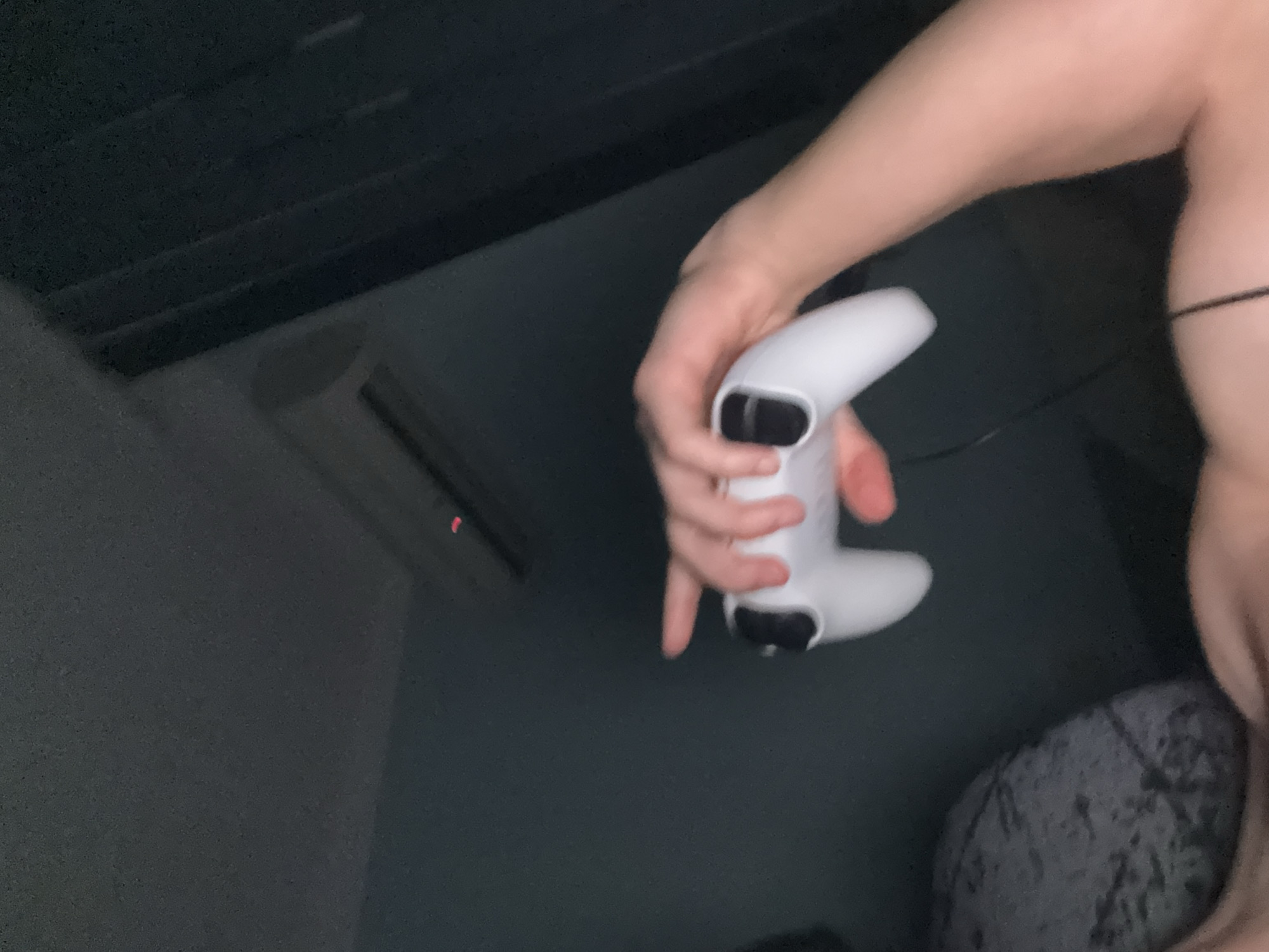 A pale-skinned person holds a white PlayStation 5 controller in their left hand. The background features a dark gray surface with a horizontal ribbed texture, possibly a wall or paneling, and a dark gray wall with an air conditioning unit on it. Part of a gray patterned pillow or blanket is visible on the right side of the image. A black wire extends from the controller. The location is not discernible from the image, so providing the city of Buhovo, Bulgaria is impossible.