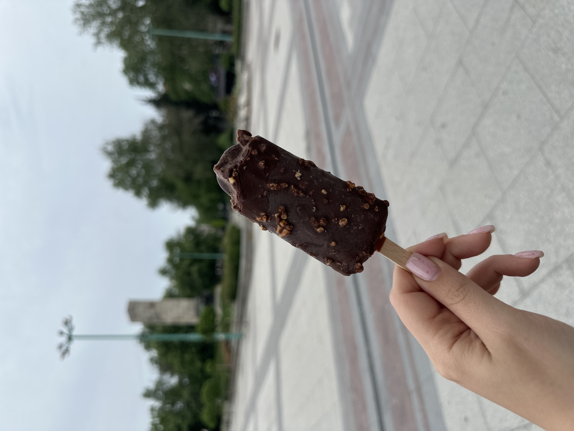 A person's right hand is holding a chocolate-covered ice cream bar on a wooden stick in front of a pedestrian plaza. The ice cream bar has nuts on the chocolate coating. The person has painted fingernails. The plaza is paved with light-colored stone or concrete tiles arranged in a geometric pattern. In the background are trees, a lamppost, and a monument. The location is in Vidin, Bulgaria.