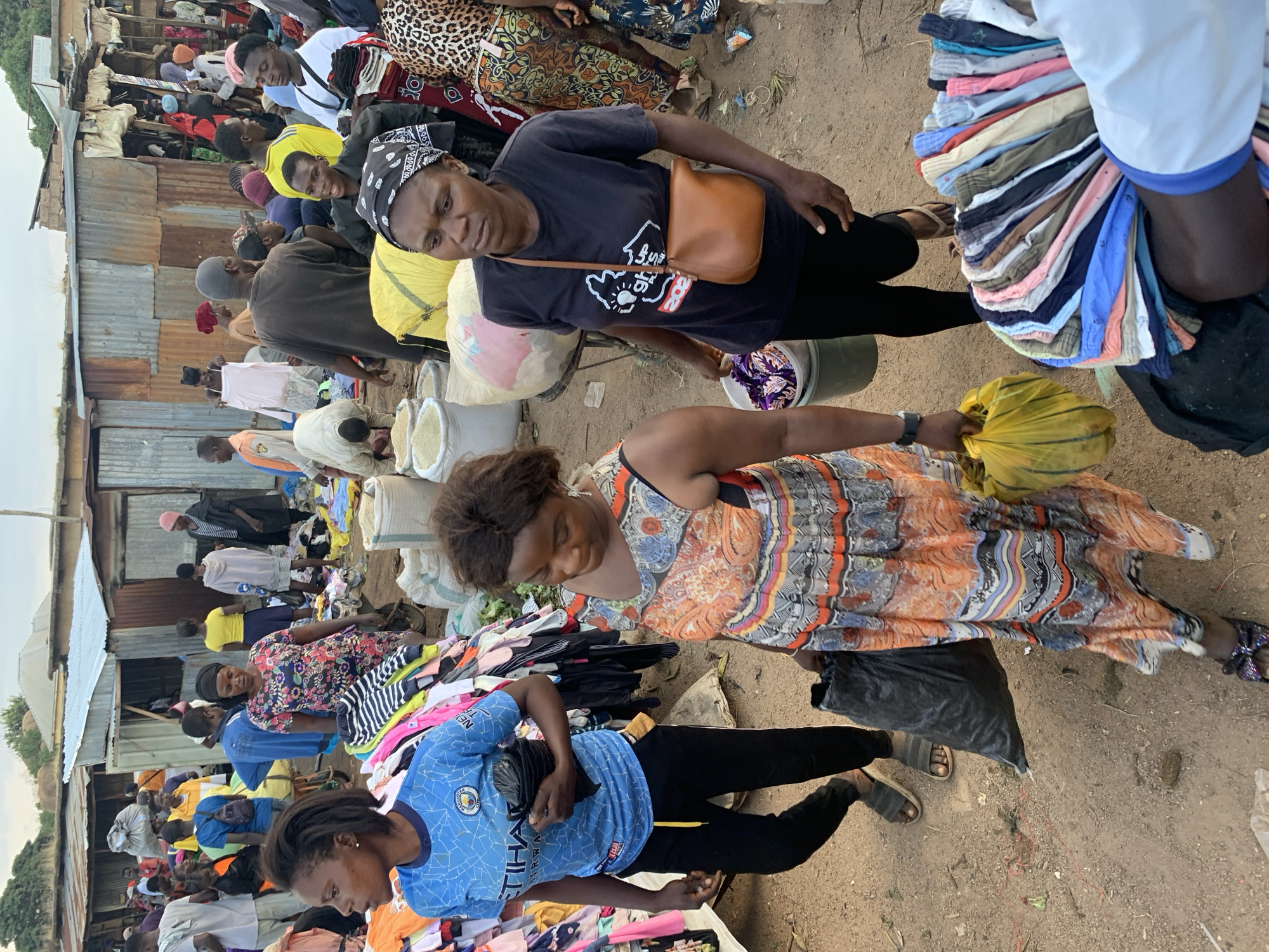 The image depicts a bustling outdoor market scene in Gura Top, Nigeria. In the foreground, two women are prominently featured. The woman on the left, wearing a blue Manchester City football jersey and black trousers, holds a black plastic bag in her right hand. To her right, another woman is seen from the side, wearing a colorful, sleeveless, floor-length patterned dress and carrying a black plastic bag in her left hand and a yellow plastic bag in her right. She has a brown crossbody bag slung over her shoulder.
The background reveals a crowded marketplace with numerous people engaged in various activities. Stalls constructed from corrugated metal and wood are visible, displaying a variety of goods, including stacks of clothing and large sacks, possibly containing grains or other commodities. The ground is dusty and uneven, characteristic of an outdoor market. The overall atmosphere is one of active commerce and community gathering. Several other individuals, men and women of various ages, are scattered throughout the scene, some interacting with stalls, others walking, and some conversing. The setting suggests a typical day at a local market in Nigeria.