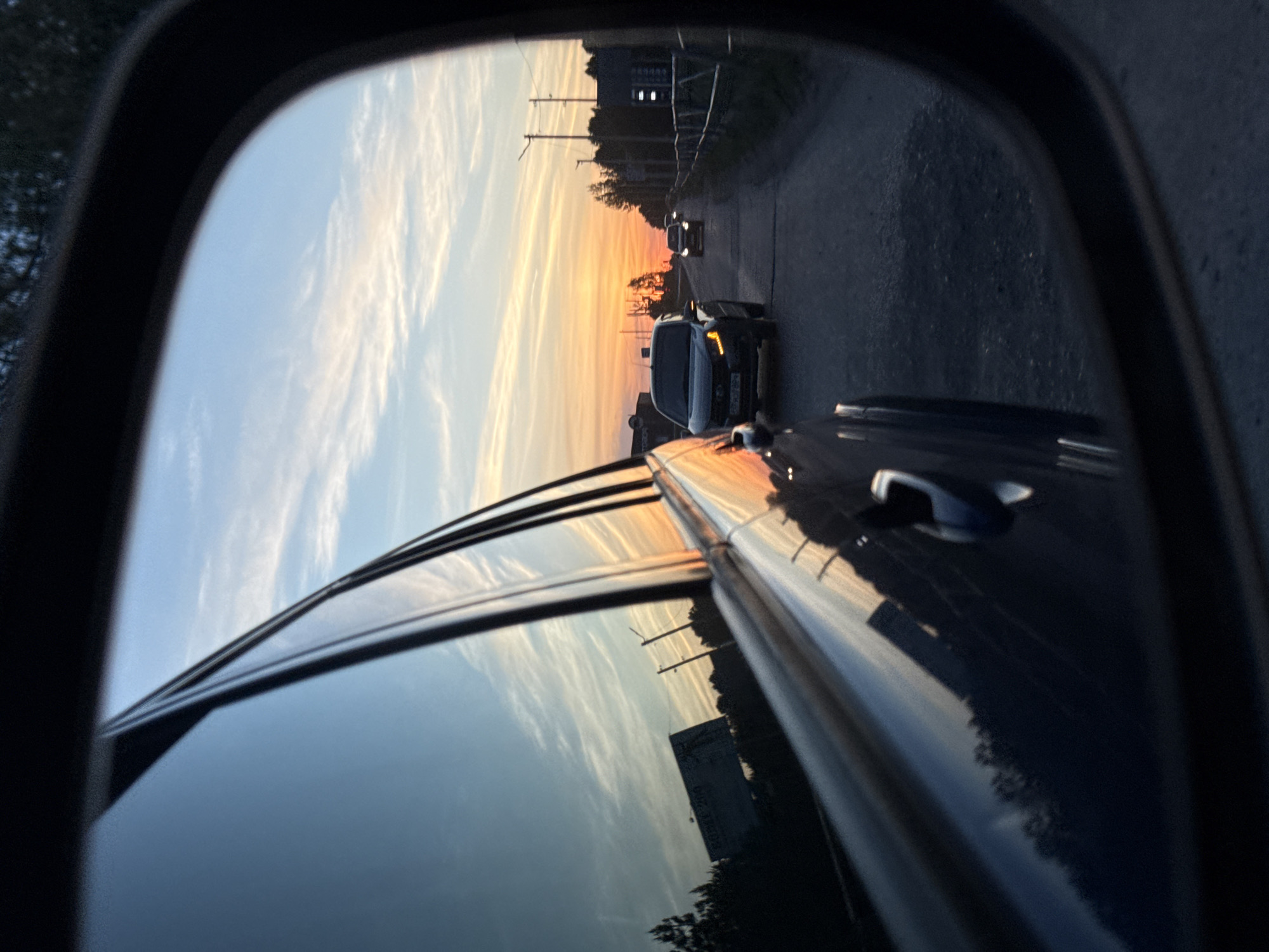 The image shows a view in a car's side mirror, reflecting a road at sunset. Another car with illuminated headlights is visible in the mirror, appearing closer than a vehicle further ahead on the road. Power lines and trees line the roadside. The sky is a mix of blue and orange hues from the setting sun, which is also reflected in the car door's surface along with the sky. The location could be near Chaikovsky, Russia given the user's description.