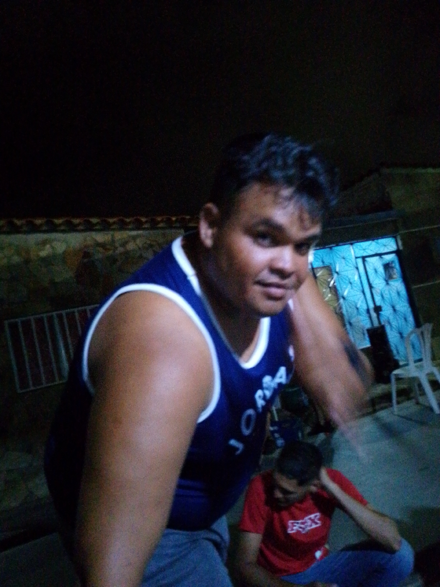 The image depicts an outdoor scene at night, likely in Juan Griego, Venezuela, given the context provided by the user's request. Two individuals are present. The primary subject, in the foreground and left of the frame, is a man wearing a blue sleeveless shirt with white trim that has "JORDAN" in white lettering. He is looking towards the camera, resting his head on his hand. His skin tone is medium to dark, and he has dark hair.
In the background, to the right and slightly further back, a second individual is seated on the ground, wearing a red t-shirt with a white logo. This person is also looking downwards, with their hand near their head. The environment appears to be a paved or concrete area, with a building visible in the background. The building has a patterned blue door or gate and a white plastic chair is situated to the right of the door. There is also a striped metal grate-like structure visible to the left of the main subject. The lighting suggests artificial light sources, common for nighttime outdoor settings.