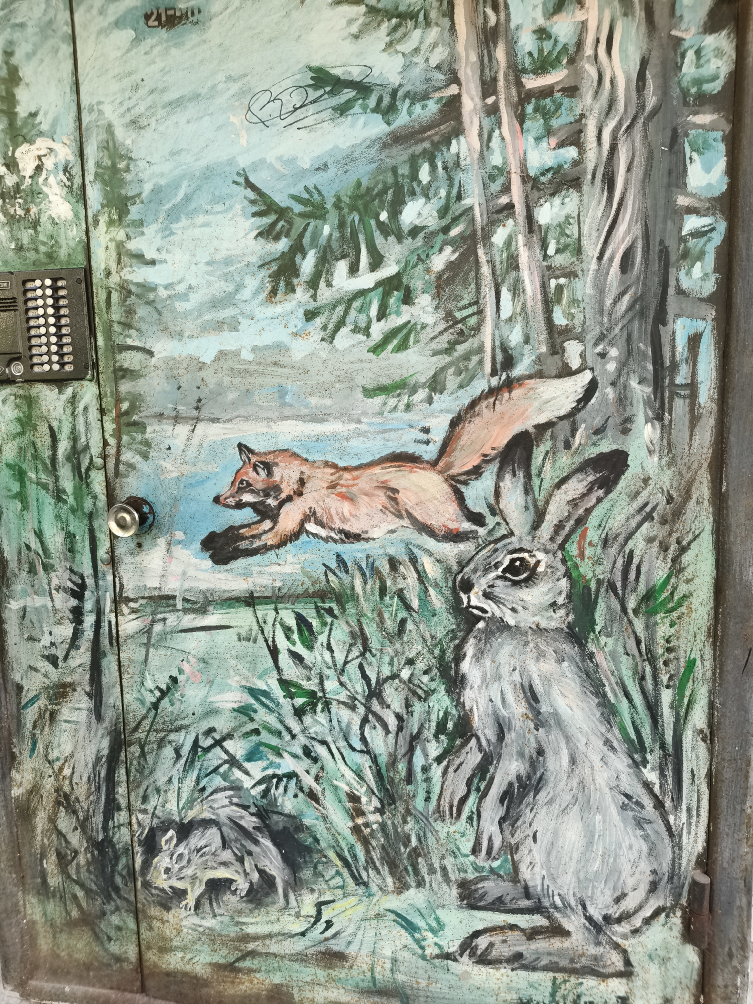 The image is a painting on a metal surface, possibly a door or utility box, depicting a forest scene with animals. A fox is depicted mid-jump across the center of the painting. To the right of the fox, a rabbit is shown in a crouched position, facing left. In the lower left corner, a small squirrel-like creature is nestled among foliage. The background features trees and a light blue sky. The painting style is loose and impressionistic, with visible brushstrokes. On the left side of the painting, a black intercom system with numbered buttons is mounted on the surface. A silver doorknob is positioned to the left of the fox. The overall condition of the painted surface appears worn, with some rust and chipped paint visible, suggesting it is an outdoor installation. The location is identified as Chaikovsky, Russia.