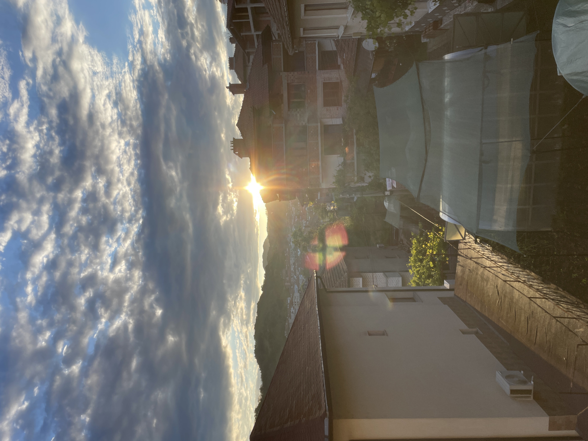The image presents a sunrise or sunset over the city of Zlatograd, Bulgaria, viewed from an elevated perspective. The sky features a mix of scattered altocumulus and stratocumulus clouds, with bright white edges illuminated by the sun, and patches of clear blue sky visible, notably in the upper right. The sun is positioned low on the horizon, slightly left of the image center, emitting a strong starburst effect and lens flare, casting warm, golden light across the scene.
In the background, rolling hills covered with dense green trees and vegetation form the natural setting for the city. Numerous buildings with red-tiled roofs are visible across the urban landscape of Zlatograd, situated on the slopes.
In the immediate foreground on the left, a light-colored building with a prominent dark red hip roof is visible, featuring multiple small rectangular windows and an outdoor air conditioning unit mounted on its lower wall. A paved walkway or ramp with a dark metal railing extends from this building towards the lower right. To the right of the sun, a multi-story building appears to be under construction or renovation, characterized by exposed brickwork, multiple balconies, a red-tiled roof, and several chimneys. Additional red-roofed buildings are visible further into the mid-ground.
The bottom portion of the image reveals several ground-level structures resembling greenhouses, covered with green mesh netting. Various green foliage and a white satellite dish mounted on a wall are also present in this area. No individuals are depicted in the scene.