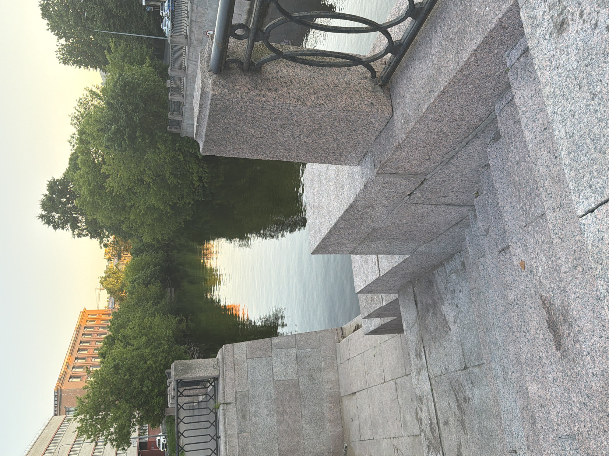 The image provides a detailed view of the Griboyedov Canal in Saint Petersburg, Russia, captured during the golden hour. In the foreground, a wide, grey granite staircase descends towards the canal, exhibiting minor wear and staining. To the right of the stairs, a robust granite pillar supports a black wrought-iron railing featuring interconnected oval patterns. Another section of the granite embankment wall is visible to the left.
The central feature is the calm, dark green water of the Griboyedov Canal, which strongly reflects the warm, golden light of the sky and the dense, green foliage lining both banks. The canal's edges are heavily treed with mature deciduous trees, whose canopies create a verdant backdrop.
On the left bank, beyond the trees, two multi-story buildings are prominent. The building further left is a large, light grey structure with a grid of windows. Adjacent to it, a reddish-brown building with a classical or monumental style is illuminated by the low sun; this building is identifiable as the main edifice of the St. Petersburg State University of Economics. A white car is partially visible near the base of the grey building, and a faint construction crane appears in the distant background. On the right bank, glimpses of a road with parked cars and a black streetlamp post are discernible through the dense foliage. The sky is a clear, pale yellow-orange gradient, characteristic of either sunrise or sunset. No people are visible in the serene urban landscape.