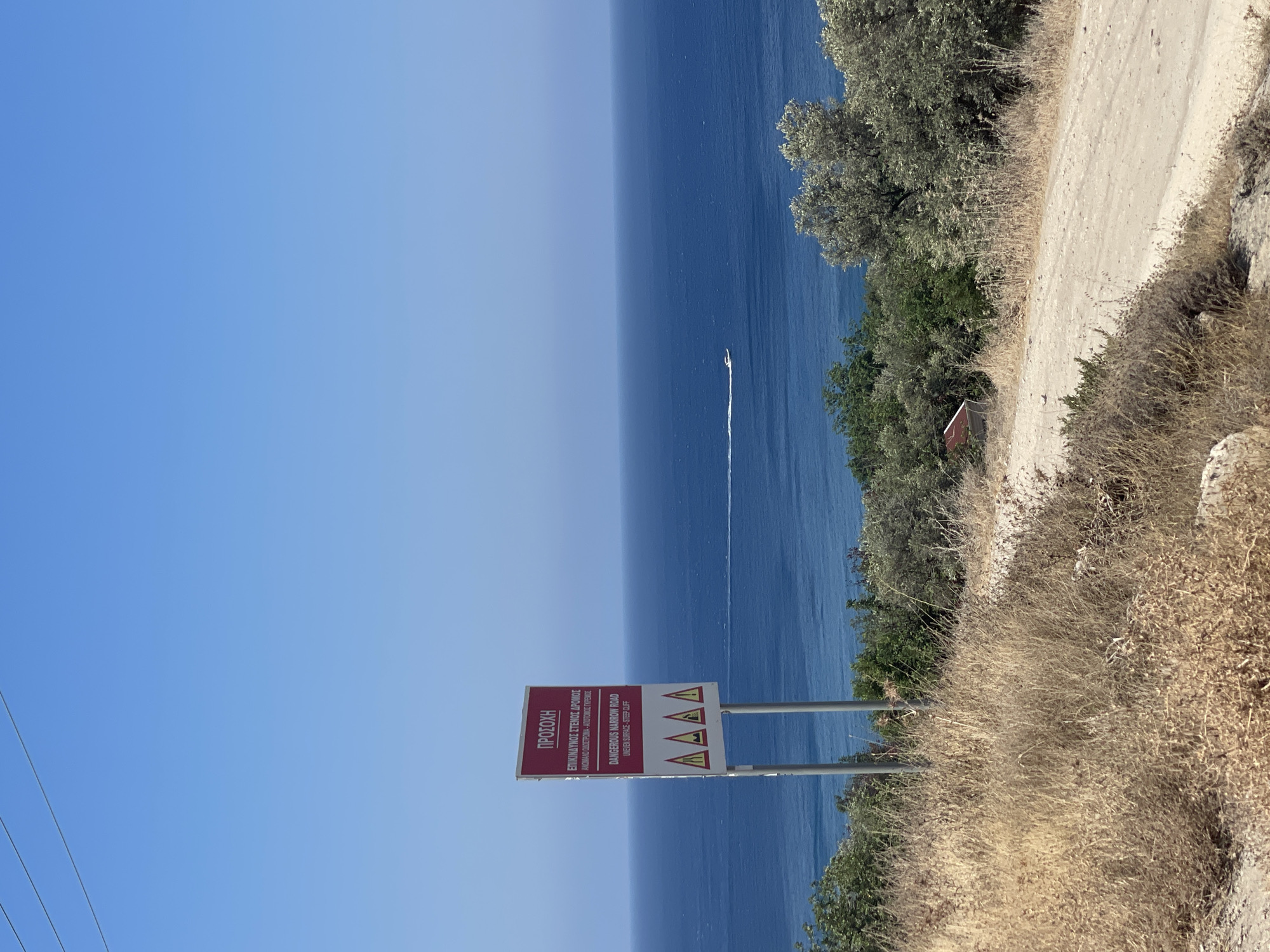 The image depicts a signpost at Neo Chorio, Cyprus, overlooking a bright blue sea. The sign, predominantly red and white, warns of a "DANGEROUS NARROW ROAD" with "UNEVEN SURFACE - STEEP CLIFF." Below this text are four triangular warning symbols, each displaying a red border and a white interior, likely representing hazards.
The signpost is situated on a dry, grassy hillside, with a dirt road curving into the foreground. To the right of the sign, a small white boat is visible on the tranquil sea, leaving a faint white wake. Lush green trees and shrubs dot the landscape, contrasting with the arid foreground. The sky is a clear, cloudless blue, indicative of a sunny day. The overall scene suggests a remote, coastal location in Cyprus, likely frequented by vehicles due to the road and warning sign.