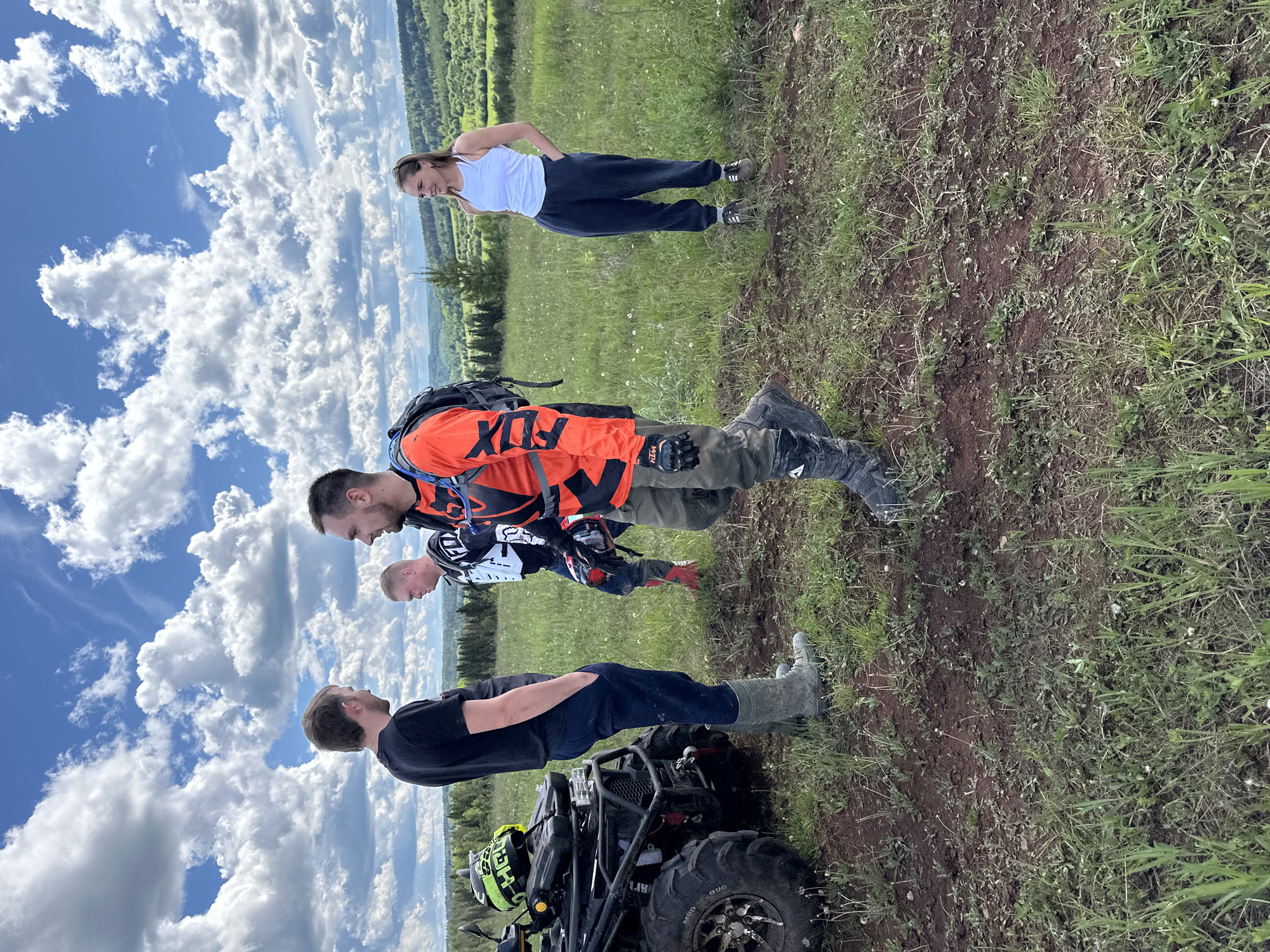 The image shows three men and a woman standing on a grassy hill with a clear sky featuring many white clouds above. The location is near Ilyinsky, Russia. The first man is wearing a black t-shirt and navy pants, along with boots. He stands beside an ATV. The second man is wearing an orange and black motocross shirt with the word "FOX" on it, green pants, and motocross boots. He is carrying a helmet. The third man is wearing a motocross shirt. The woman is wearing a white tank top and dark pants, with hands on her hips. The background includes a green field and trees.