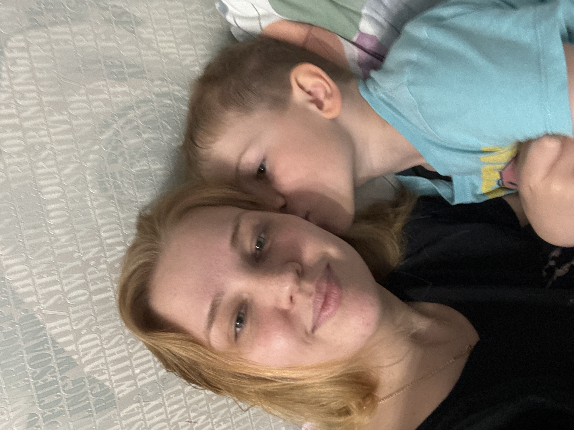 A woman with blonde hair is lying down with a young boy in a light blue shirt with a graphic on it, who is leaning close to her face. They are on a patterned bed sheet with what appears to be text embossed on it. The scene is indoors. The city of Rusanovo, Russia, is not determinable from the image.