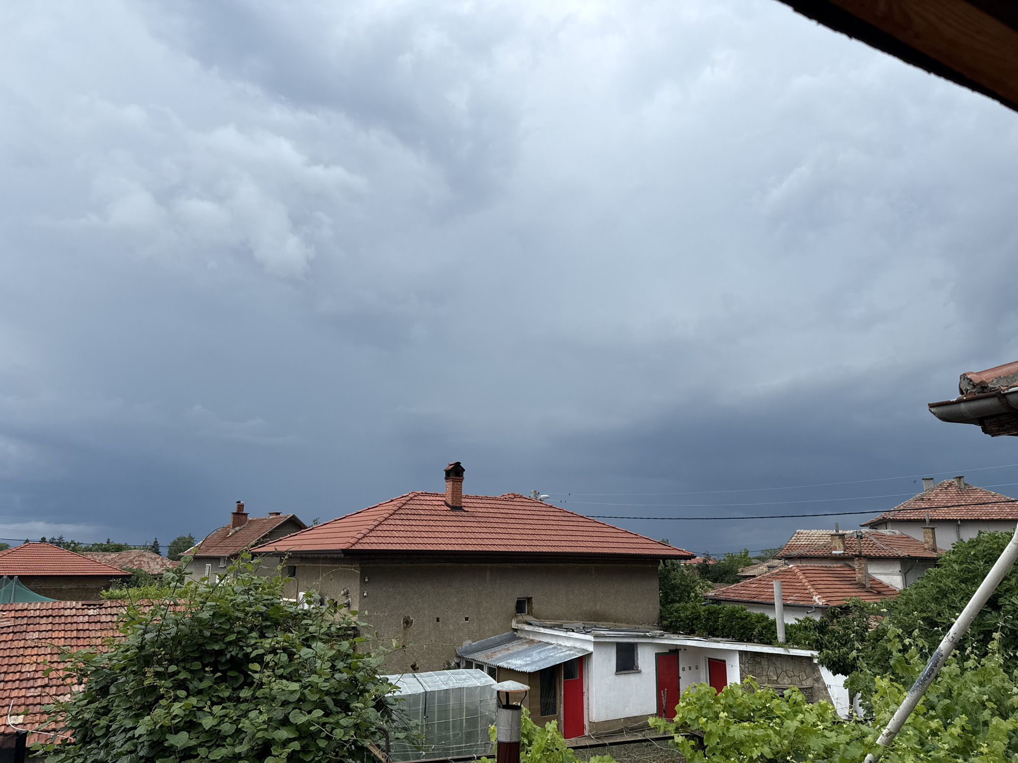 The image presents an overcast daytime view of a village, identified as Hadzhidimitrovo, Bulgaria, seen from an elevated perspective. The dominant feature is a vast, heavy, dark grey sky filled with dense, low-hanging clouds, strongly suggesting an impending storm or recent adverse weather.
Below the dramatic sky, a cluster of houses with characteristic terracotta red tile roofs is visible, interspersed with abundant green trees and bushes. The architecture includes buildings with stuccoed walls, some appearing in neutral tones like beige or grey. Several chimneys protrude from the rooftops, particularly on a prominent two-story house in the midground. This house features a large, simple facade and a well-maintained red tile roof. Adjacent to it, a smaller, white auxiliary building with two distinct red doors and a corrugated metal roof stands out. A greenhouse is also partially visible near the lower-left corner.
Lush green foliage, including what appears to be grapevines, fills the lower and mid-sections of the frame, obscuring some of the structures. Power lines stretch horizontally across the midground, connecting unseen poles. The scene is static, with no human activity or visible text present. The overall mood conveyed by the dark sky is one of quiet anticipation or a somber atmosphere.