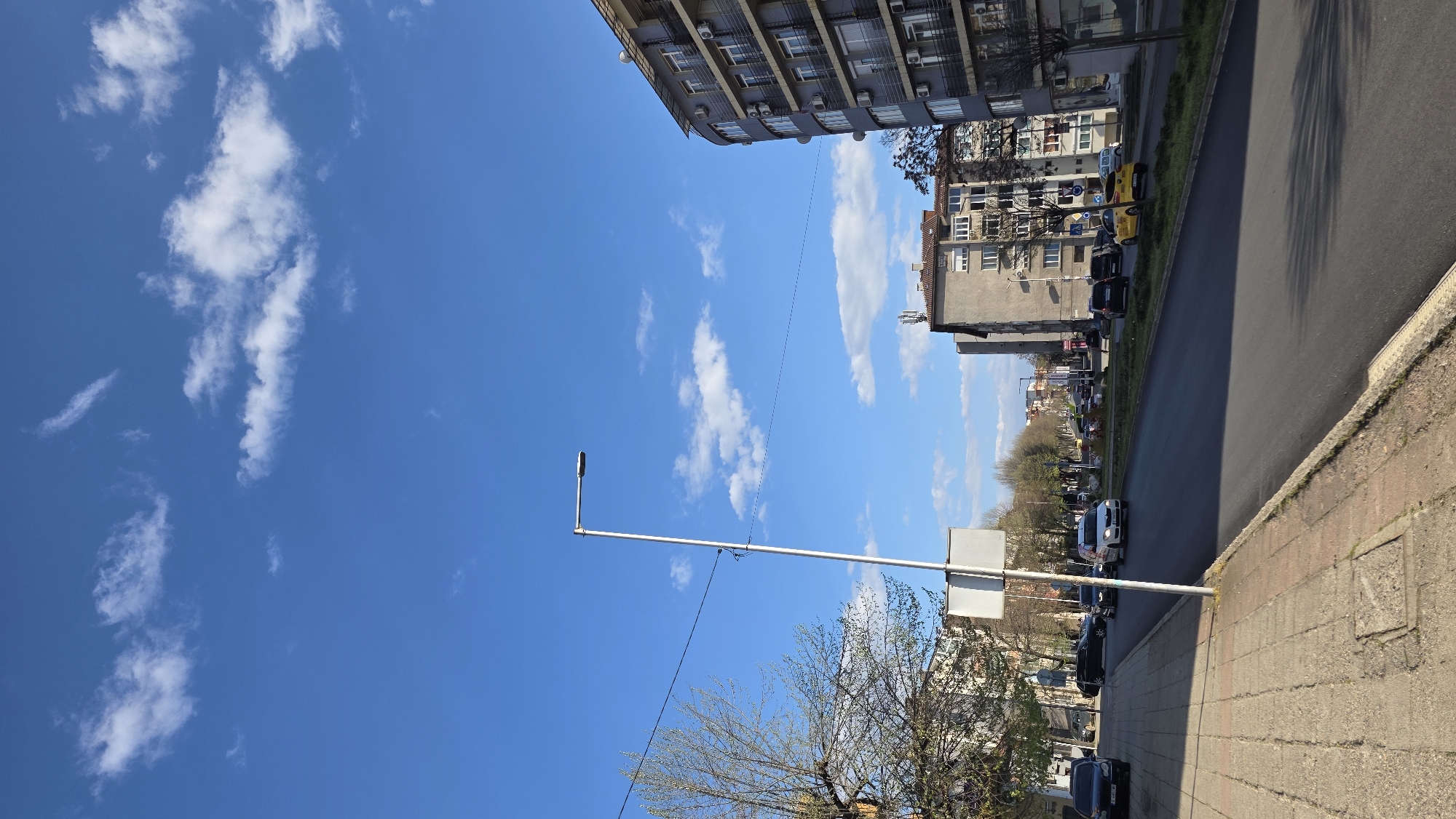 The image depicts a street scene in Blagoevgrad, Bulgaria, on a sunny day. The sky is blue with scattered white clouds. Buildings line the street, including a taller apartment building with air conditioning units and a shorter, older building with windows and balconies. Cars are parked along the side of the road, and there are trees with budding leaves. A lamppost stands tall against the sky. The road is paved, and a sidewalk runs along the edge.