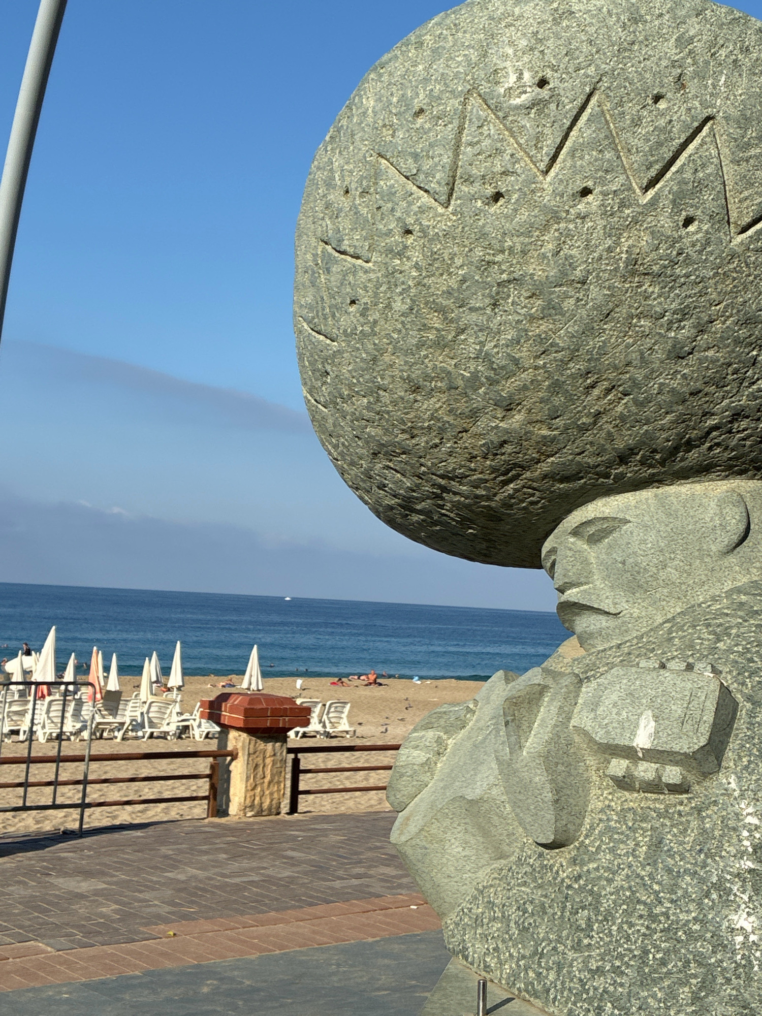 In Alanya, Turkey, a stone sculpture of a figure carrying a large, rough-hewn sphere on its head stands prominently in the foreground. The sculpture's face is rendered in a simplified, stylized manner, with the figure appearing to be wearing some sort of garment. Behind the sculpture, a sandy beach is visible, lined with rows of empty white lounge chairs and umbrellas. The sea stretches out to the horizon, with a clear blue sky above and a thin line of clouds. A few distant boats can be seen on the water. The beach area appears to be a public space, possibly a resort or park.