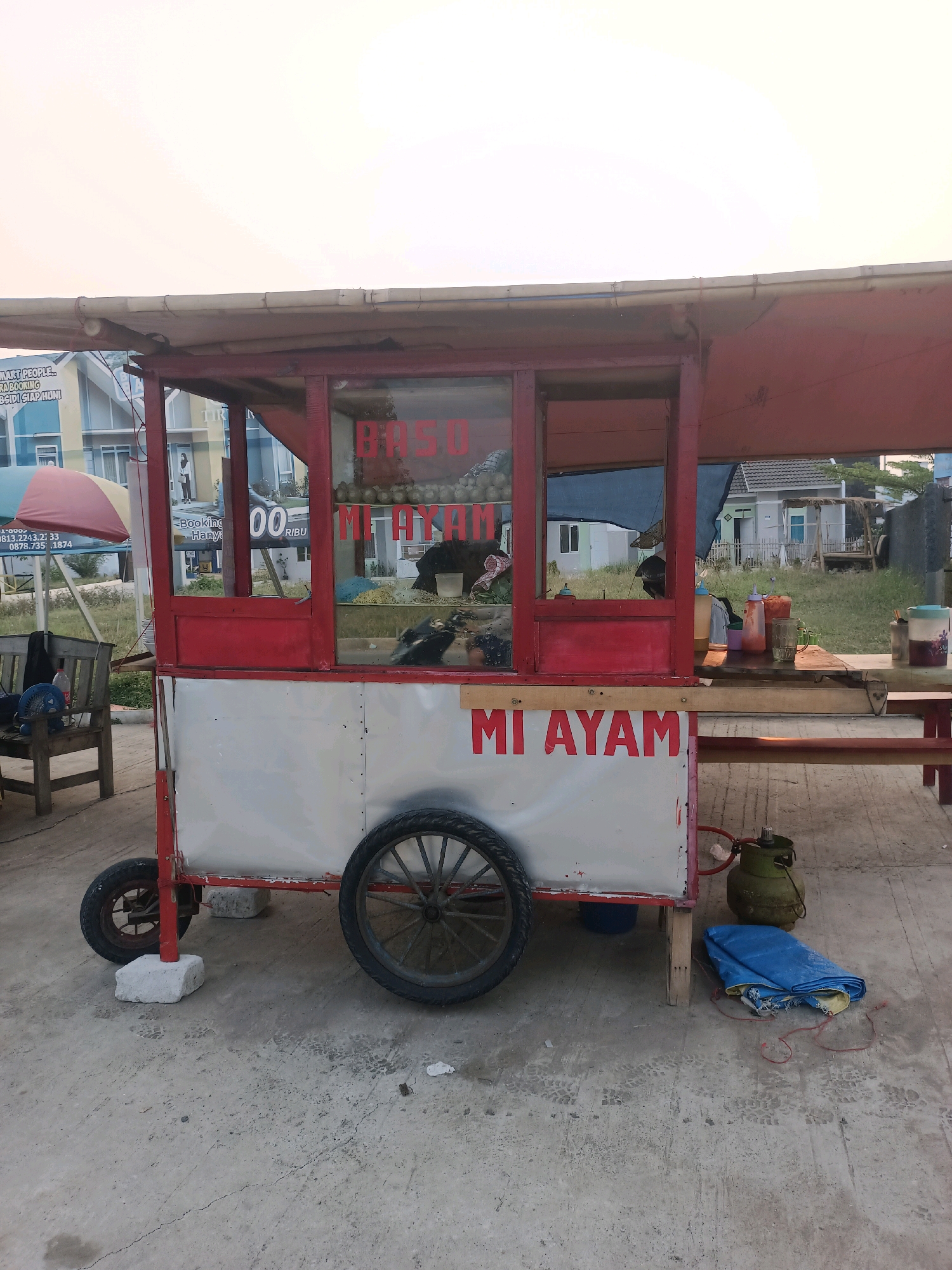 The image shows a food cart selling "Mi Ayam" (chicken noodles) and "Baso" (meatballs) parked on a concrete surface in Karawang, Indonesia. The cart is red and white, with a red awning. There are two large wheels on the cart, and it is propped up by two concrete blocks. In the background, there are buildings and some greenery. A table with condiments and utensils is set up next to the cart. A blue tarp and a green gas tank are on the ground near the cart. The sky is bright and appears to be either sunrise or sunset.