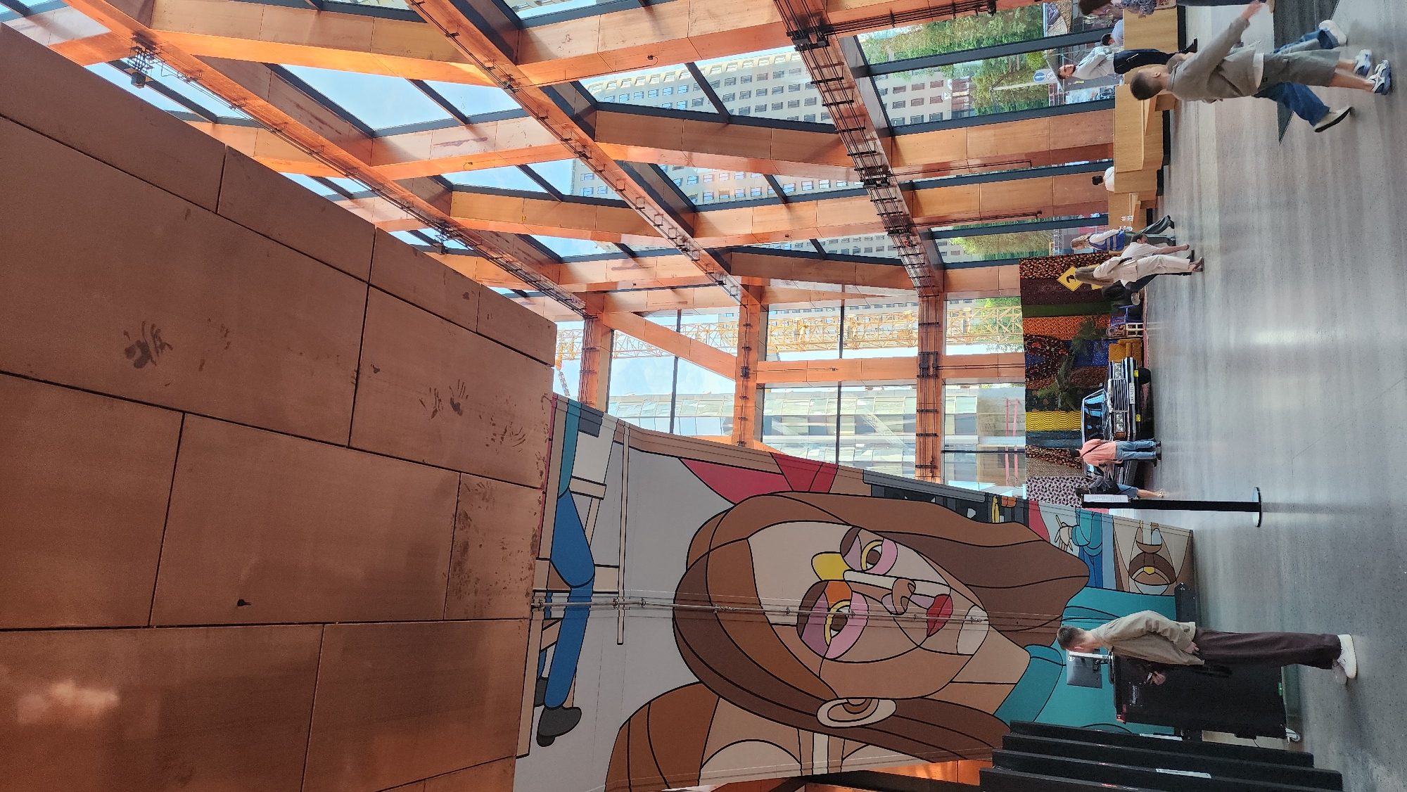 The image shows the interior of a building with a striking architectural design, presumably a museum or public space. The most notable feature is the intricate wooden beam structure forming the ceiling, interspersed with glass panels that allow natural light to flood the space. The walls are covered with large, rectangular tiles, possibly made of wood or a similar material. A large, colorful mural of a stylized face dominates the lower portion of the image.
Several people are present in the scene, appearing to be visitors to the building. They are seen standing and walking around, interacting with the exhibits and the space.
The presence of a building under construction visible through the glass ceiling suggests the building is in a city environment.
There is no reliable information to determine whether or not the image originates in the city of Moscow, Russia.