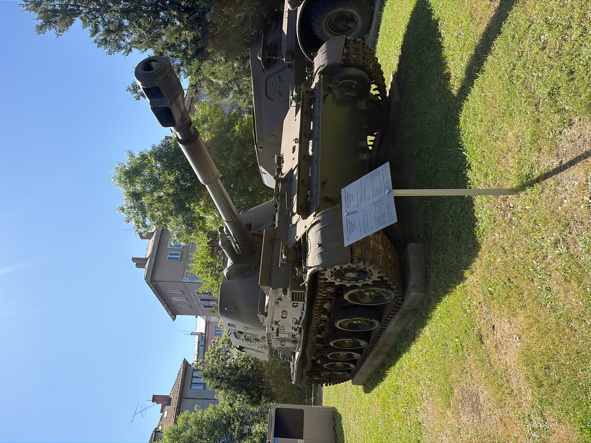 The image captures an outdoor display of a military vehicle, likely a self-propelled howitzer, on a sunny day. The vehicle, painted in a drab olive green, is positioned on a concrete base within a grassy area. It features a long barrel extending forward and a tracked chassis with multiple road wheels. Adjacent to the vehicle, a sign with information in Cyrillic script stands on a metal pole.
In the background, typical urban architecture is visible. These are multi-story buildings with tiled roofs and windows, suggesting a residential or public area. Lush green trees frame the scene, and the bright blue sky with some sun glare indicates clear weather. To the left of the main subject, a modern, light-colored structure, possibly a kiosk or information booth, is partially visible. Another military vehicle, a wheeled one, is partially visible behind and to the right of the main subject.
The overall setting appears to be a museum or memorial park in an urban environment. Based on the Cyrillic text on the sign, specifically mentioning "Sofia, Bulgaria" and "National Museum of Military History", this location is identified as the National Museum of Military History in Sofia, Bulgaria. The sign itself provides technical specifications for the displayed "2S23 Nona-SVK Self-Propelled Howitzer".