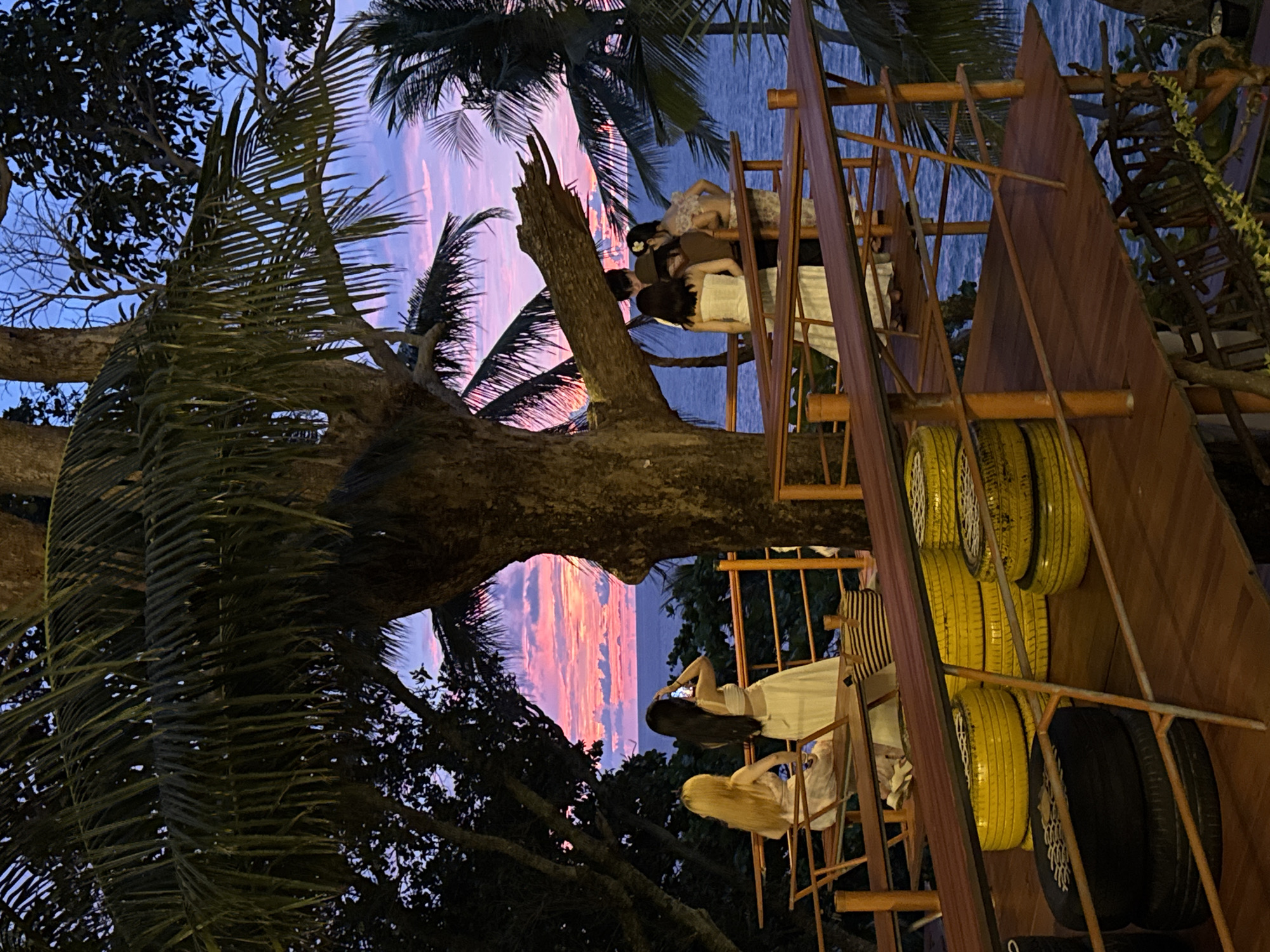 In Karon, Thailand, a group of individuals gathers on a wooden deck overlooking the ocean at sunset. The sky displays vibrant hues of pink, purple, and blue as the sun dips below the horizon. Palm trees frame the scene, their fronds casting shadows on the deck. Several people are visible: two women are seated on a bench in the foreground, one with blonde hair and the other with long black hair. The woman with black hair is holding a phone up as if taking a picture. Further back, a group of four people are standing, appearing to converse or observe the sunset. The deck itself is constructed from wooden planks and features a railing. In the lower portion of the image, stacks of yellow and black tires are used as seating or decorative elements. The ocean stretches out to the horizon, its surface reflecting the colors of the sky.