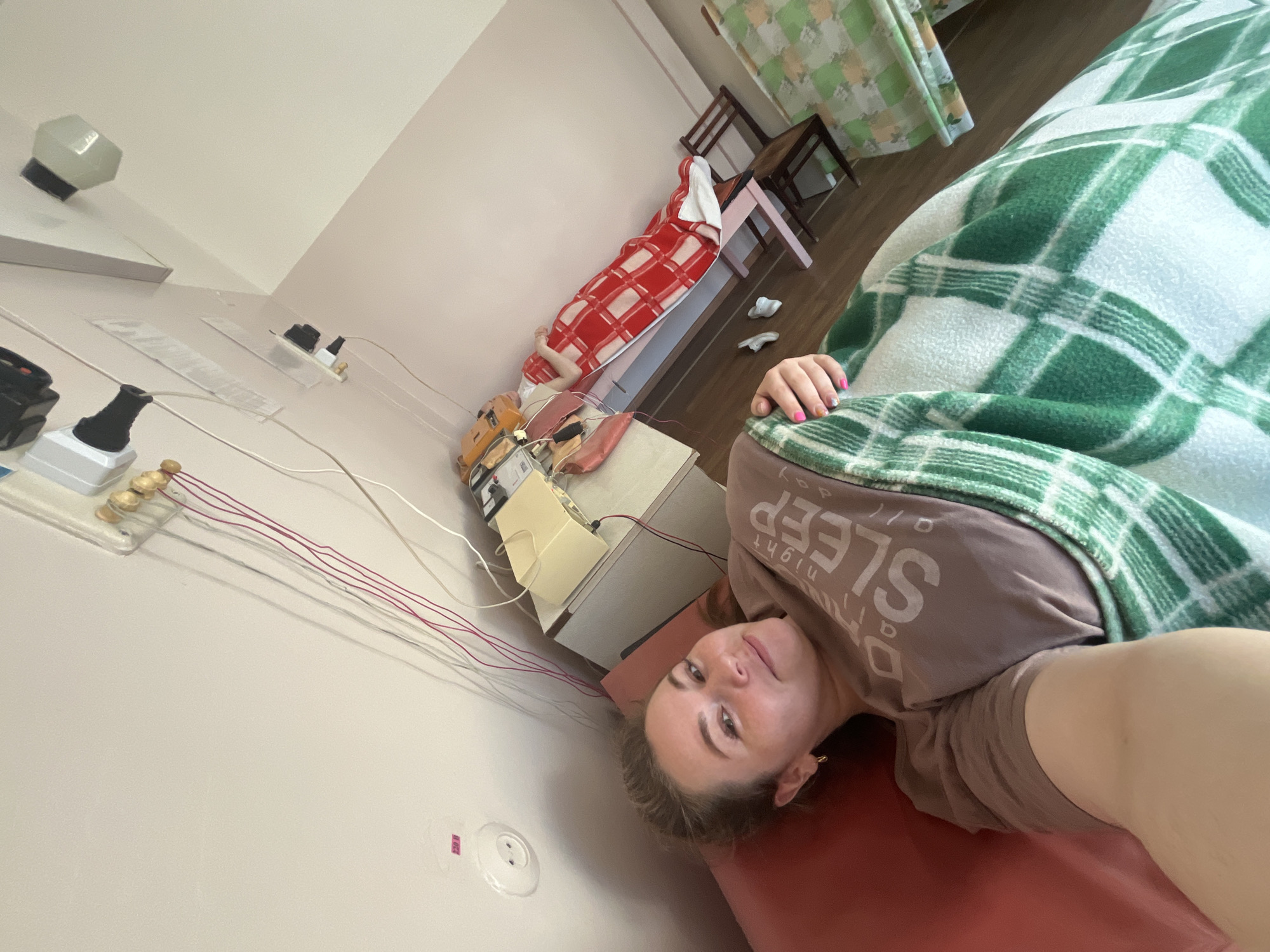A woman is lying on a bed in a room. She is wearing a brown shirt with the words "NIGHT SLEEP" on it and is covered with a green and white checkered blanket. Her right hand is visible near the top right corner of the image. Behind her, another person lies under a red and white blanket on a separate bed, positioned against the wall. There is medical equipment on a nearby table, and wires connect it to the wall. The room has light-colored walls, and furniture is visible in the background. The image was likely taken in the city of Chaikovsky, Russia, given the context provided in the prompt.