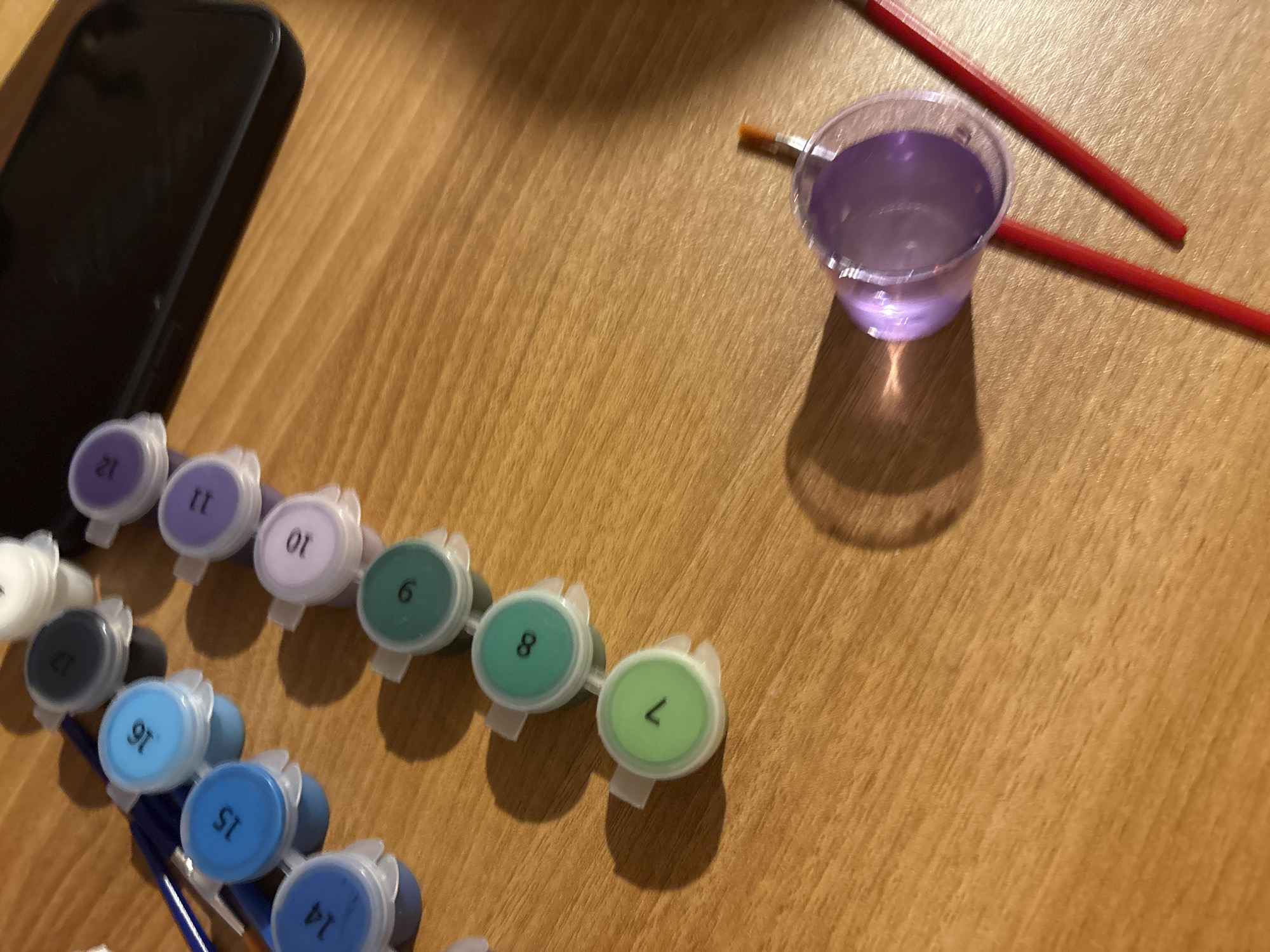 A close-up shot shows a wooden table. On the left side of the table is a black smartphone next to paint containers labeled with numbers from 6 to 21. A clear plastic cup filled with purple water sits near two red-handled paintbrushes. The environment is indoors. There is no evidence to indicate that the scene is in Plovdiv, Bulgaria.