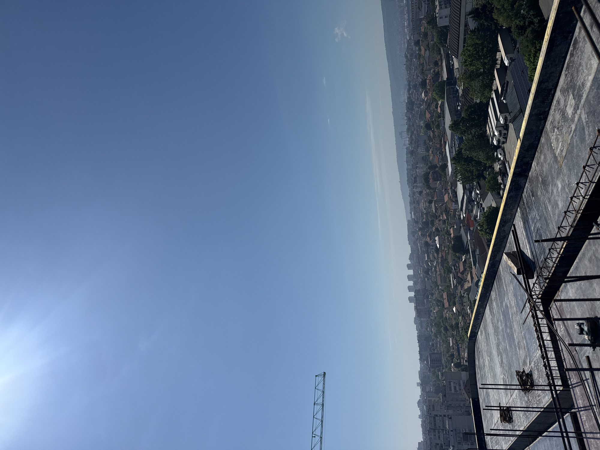 This media file presents a wide, high-angle view of a city under a clear blue sky, likely taken from a significant height, possibly a tall building or construction site. The image is oriented vertically, showing the sky dominating the upper half and the city extending towards the horizon in the lower half.
The sky is a vibrant blue with a bright lens flare from the sun visible in the upper left, indicating a sunny day. The horizon line is somewhat hazy, suggesting atmospheric distance. A vast urban landscape unfolds below, characterized by a dense array of buildings, including residential structures with red-tiled roofs, taller apartment blocks, and commercial/industrial zones with larger buildings and what appear to be parked semi-trucks. Patches of green foliage, primarily trees, are interspersed throughout the urban fabric. In the distant background, gentle hills or mountains are visible, partially obscured by haze.
In the foreground, occupying the right side of the image, is a partially constructed concrete rooftop or floor slab of a building. Exposed rebar and structural elements are prominent, indicating ongoing construction. Part of a tall metal lattice structure, resembling a crane tower, extends into the lower center of the sky, further suggesting a construction environment.
No people or specific activities are discernible. The time of day is clearly daytime, with bright sunlight. The weather is clear and sunny. Given the context, this is a view over Varna, Bulgaria, showcasing its urban development and surrounding natural landscape from above. No visible text is present in the image.