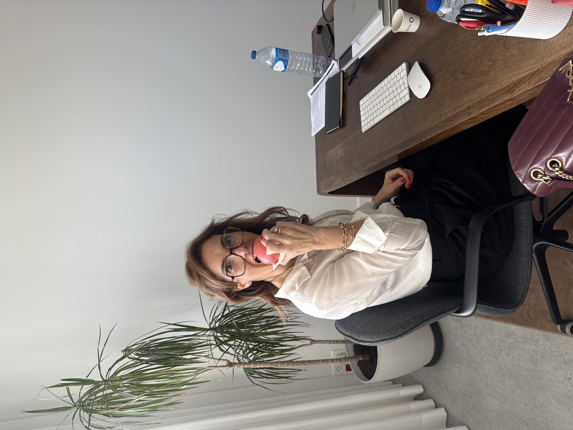 A woman with brown hair and glasses is sitting in an office chair, positioned at a wooden desk. She is wearing a white shirt and has multiple bracelets on her left wrist. She is eating an apple. On the desk, there is a laptop, keyboard, mouse, water bottle, a container with office supplies, and a handbag. Beside the desk, a large potted plant with long, green leaves is visible. The scene is likely indoors, within an office space, suggested by the furniture and equipment. The location is not discernable as being within the city of Torres Vedras, Portugal, based on the available image.