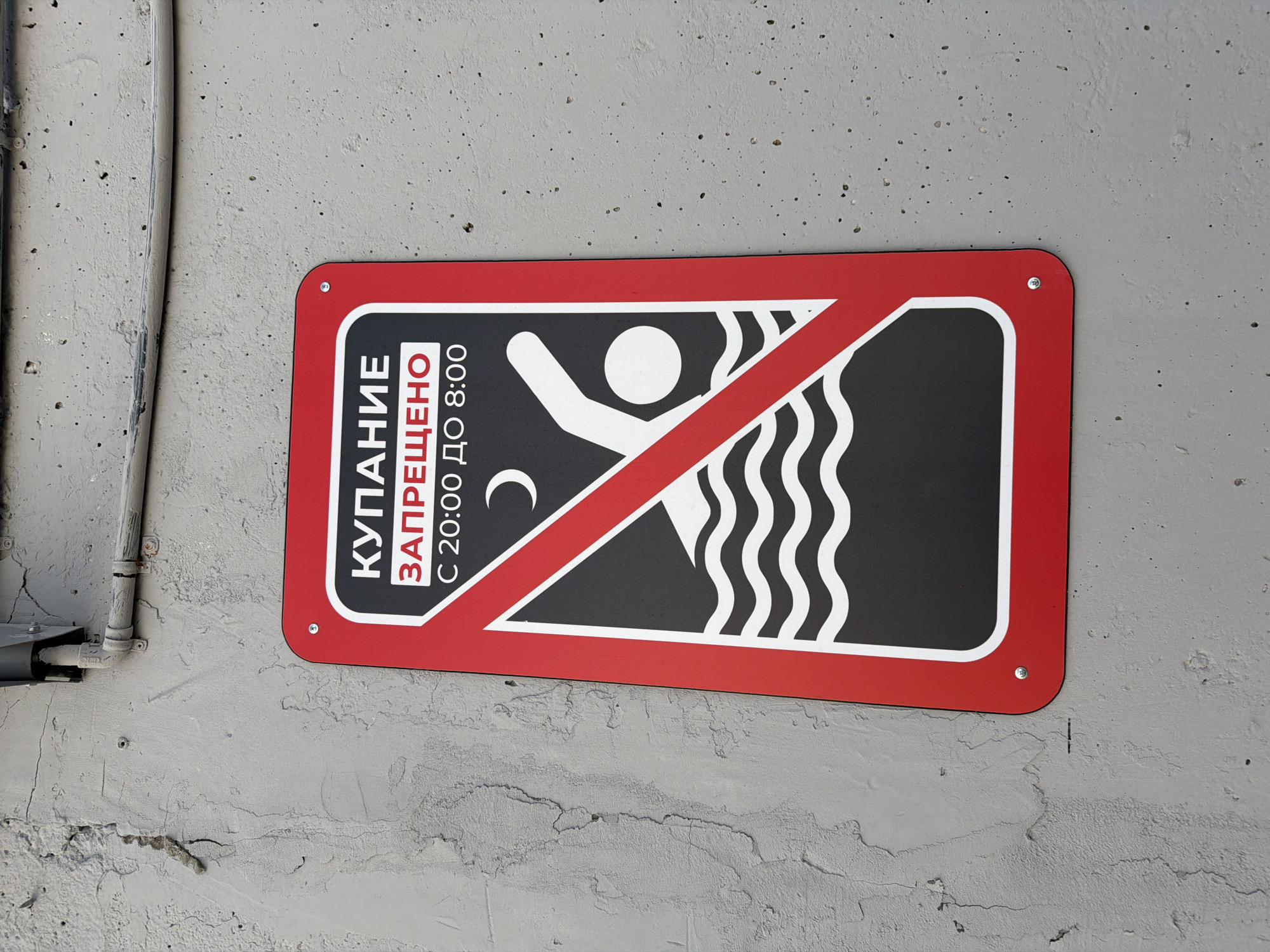 A red and white prohibition sign is affixed to a textured gray concrete wall. The sign displays Russian text that translates to "SWIMMING FORBIDDEN FROM 20:00 TO 8:00." It features a red diagonal line across a black rectangle, which contains a stylized graphic of a crescent moon, a swimmer, and wavy lines representing water. The sign is secured to the wall with visible screws. Above the sign, a metal conduit with wires runs horizontally along the wall. The wall exhibits a rough, weathered surface with visible cracks and imperfections.