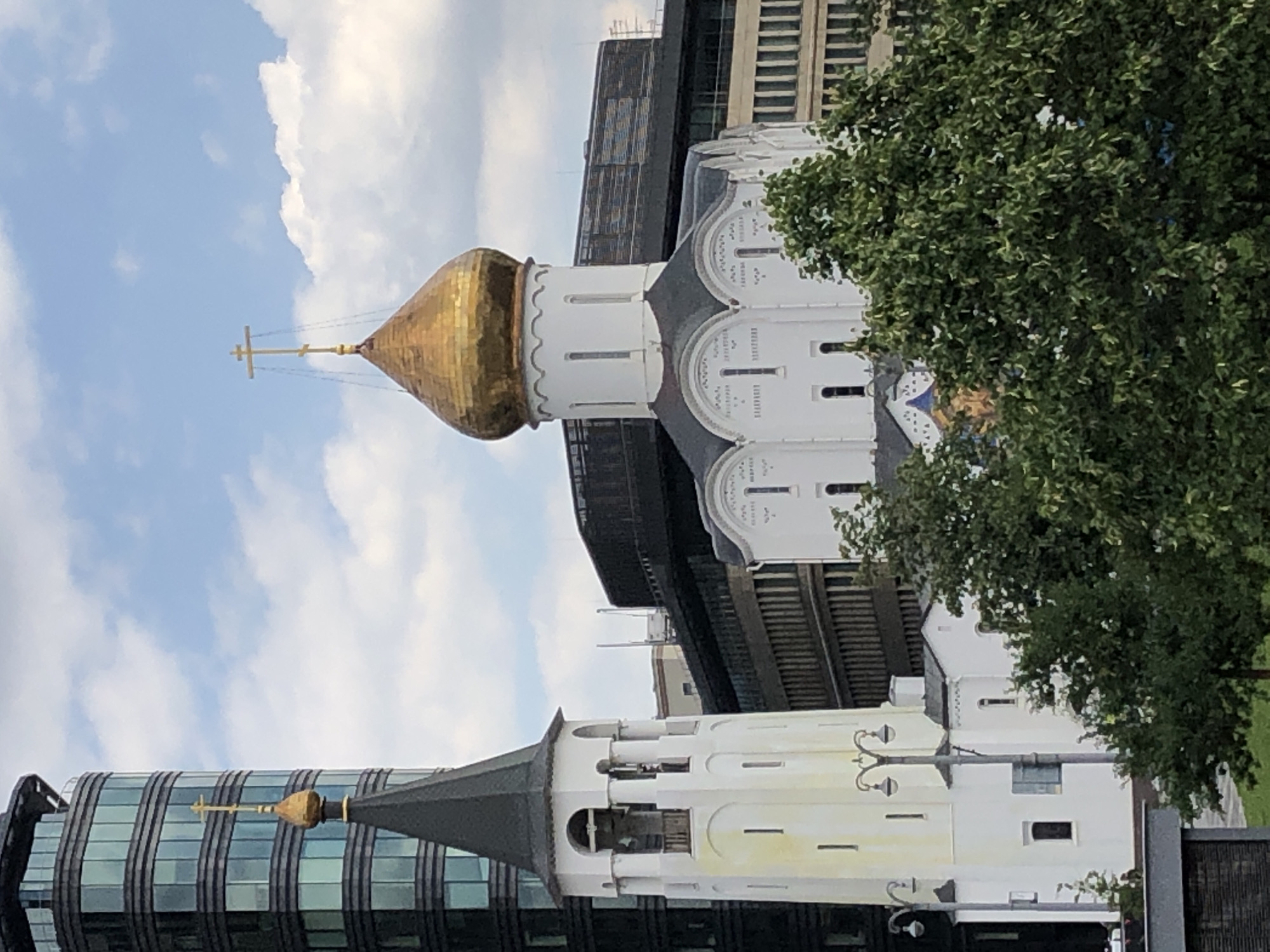 This is a photograph taken from an elevated position, looking towards a modern cityscape that includes a church and its bell tower. The modern building on the left is tall and cylindrical, with a glass facade composed of horizontal segments. To its right, a gold-domed Orthodox church stands, characterized by its white stucco exterior and intricate architectural details. Adjacent to the church is a white bell tower, slender and topped with a dark, pointed roof. The scene is framed by lush green trees in the foreground and background, with a cloudy sky above. The image appears to be taken in Moscow, Russia, as suggested by the architectural style of the church and bell tower, which are typical of Russian Orthodox churches. There are no people visible in the frame. The overall impression is one of a juxtaposition between historic religious architecture and contemporary urban development.