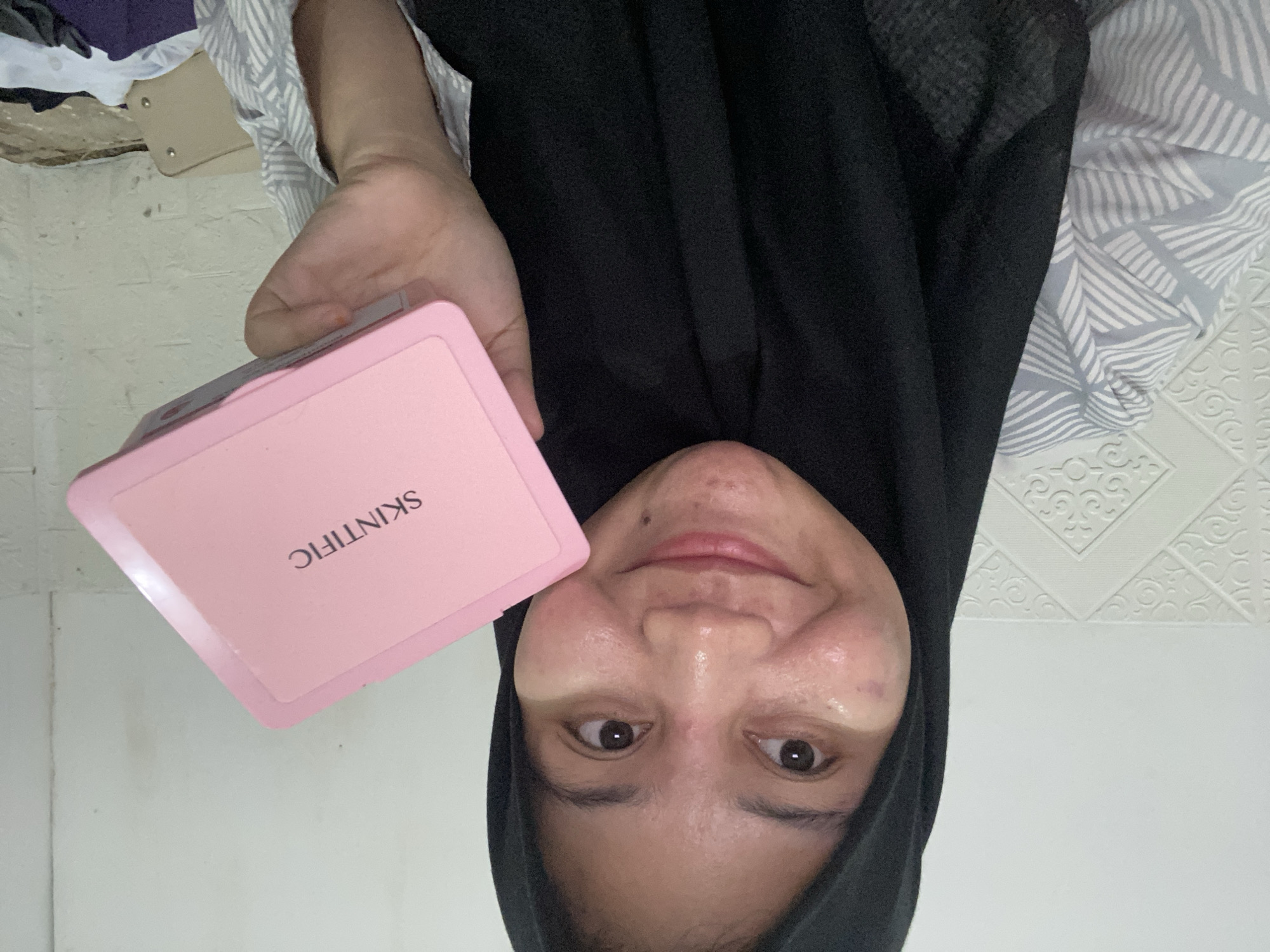 A medium shot captures a woman wearing a black hijab and a grey and white striped shirt. She is holding a pink rectangular box labeled "SKINTIFIC" in her right hand. The woman is looking at the camera with a slight smile. The background consists of a white textured wall and a patterned surface behind her left shoulder. The scene appears to be indoors.