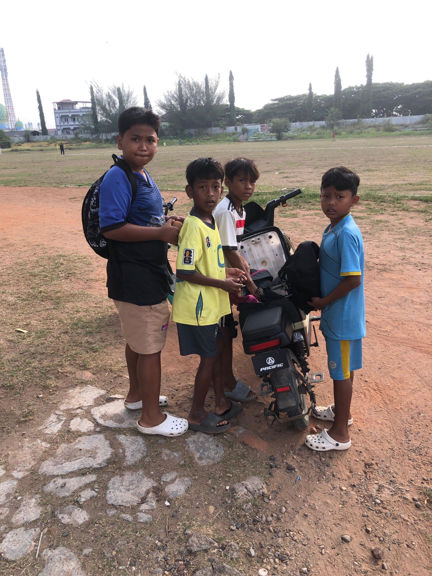 The image depicts four young boys standing around a motorcycle on a dirt and grass field, likely a sports field, in Lhokseumawe, Indonesia. The boys are casually dressed in t-shirts and shorts, with some wearing sandals or flip-flops. One boy on the left wears a blue t-shirt and black shorts, with a backpack slung over his shoulder. The second boy from the left wears a yellow t-shirt and dark shorts, holding something small in his hand. The third boy from the left wears a white and red striped t-shirt and dark shorts. The boy on the right wears a blue t-shirt and blue and yellow shorts, holding a black bag. The motorcycle is black and white with "PACIFIC" written on the rear fender. In the background, there are trees, a fence, and buildings, including what appears to be a tower. The overall lighting suggests it is daytime, possibly late afternoon, with overcast skies. The ground is a mixture of reddish-brown dirt, patches of dry grass, and some embedded stones.