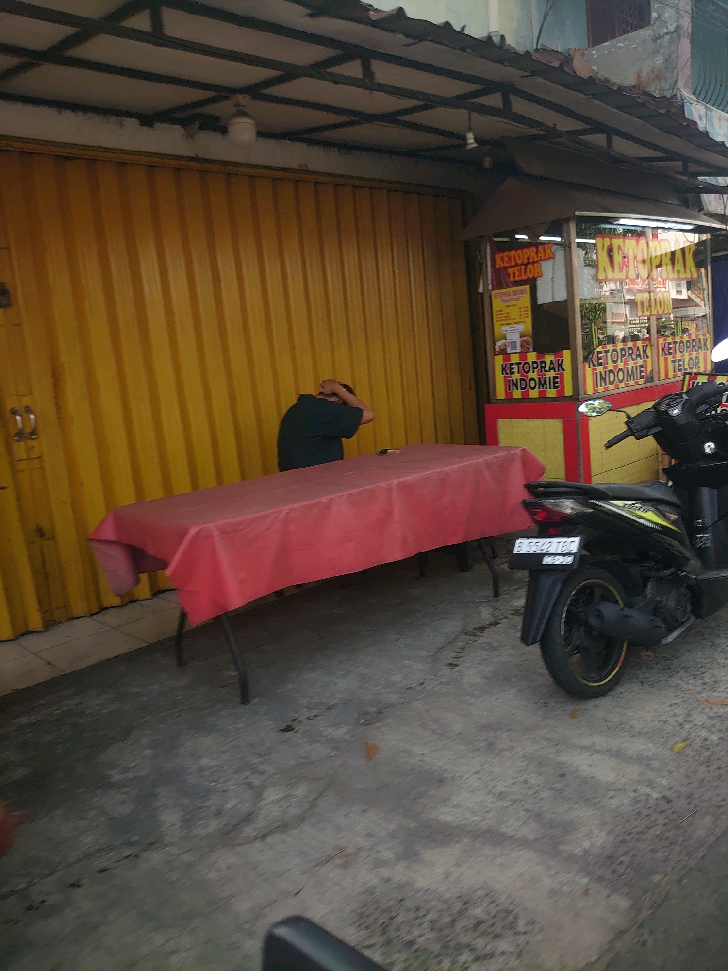 In Depok, Indonesia, a street-side food stall is captured in an image. A yellow corrugated metal gate serves as the backdrop for a long, red-clothed table. Seated at the table, a person wearing a dark shirt has their head bowed and hands over their head, suggesting distress or fatigue. To the right of the scene, a black motorcycle is parked. The stall itself is brightly decorated with signage that reads "KETOPRAK TELOR" and "KETOPRAK INDOMIE," indicating the food items sold. The overall environment appears to be an urban street.