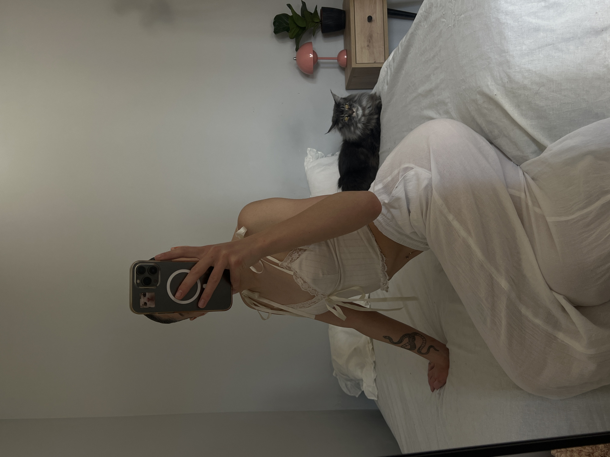The image shows a woman taking a mirror selfie in a bedroom. She is wearing a white lace bralette and white wide-leg pants, and she has a snake tattoo on her left arm. A large grey cat is lying on the bed behind her. To the right of the bed, there is a nightstand with a pink lamp and a potted plant. The room appears to be a bedroom with a light grey wall and a white bedspread. The image does not contain any identifiable landmarks or contextual clues that would allow for its location to be determined in Ternopil, Ukraine.