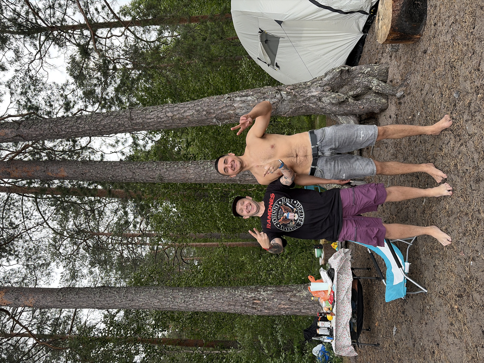 Two adult males are posing for a photo in a forest clearing near Vaskelovo, Russia. The man on the left is wearing a black t-shirt with the Ramones logo and purple shorts. He is wearing a black baseball cap and making a peace sign with his right hand. The man on the right is shirtless and wearing grey denim shorts. He is also making a peace sign with both hands. In the background, there is a table laden with food and drinks, a blue camping chair, and a white tent. The ground is covered with dirt and pine needles, and tall pine trees surround the clearing. The sky is overcast.