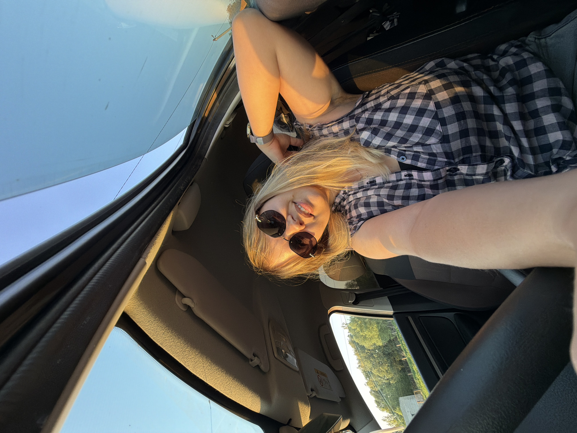 A low-angle, upside-down selfie shows a woman in a car. She wears sunglasses and a black-and-white checkered shirt. The car's interior is visible, including the dashboard, windshield, and sunroof. Outside the car window, greenery and a building are visible. The location is not discernible from the provided image.